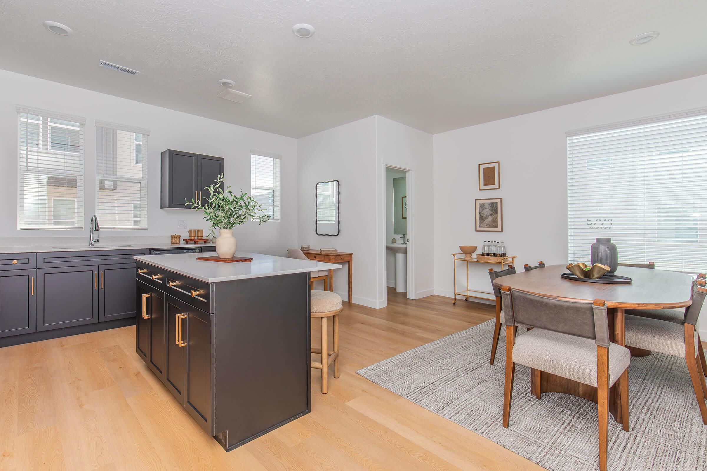 Modern kitchen and dining area featuring dark cabinetry, a large island with seating, and a dining table for six. Large windows provide natural light, and the decor includes plants, artwork, and stylish furnishings, creating a warm and inviting atmosphere.