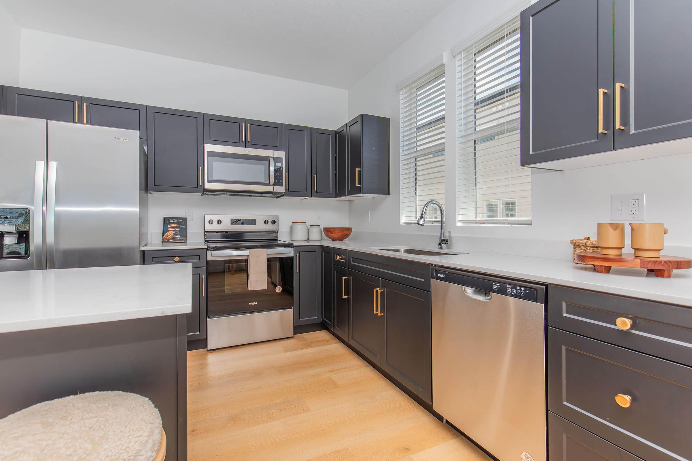Modern kitchen featuring dark cabinetry, stainless steel appliances, and a white countertop. The space includes a refrigerator, microwave, oven, and dishwasher, along with a sink and natural light from windows. Decor elements include wooden bowls and countertop accessories. Stylish and functional design.