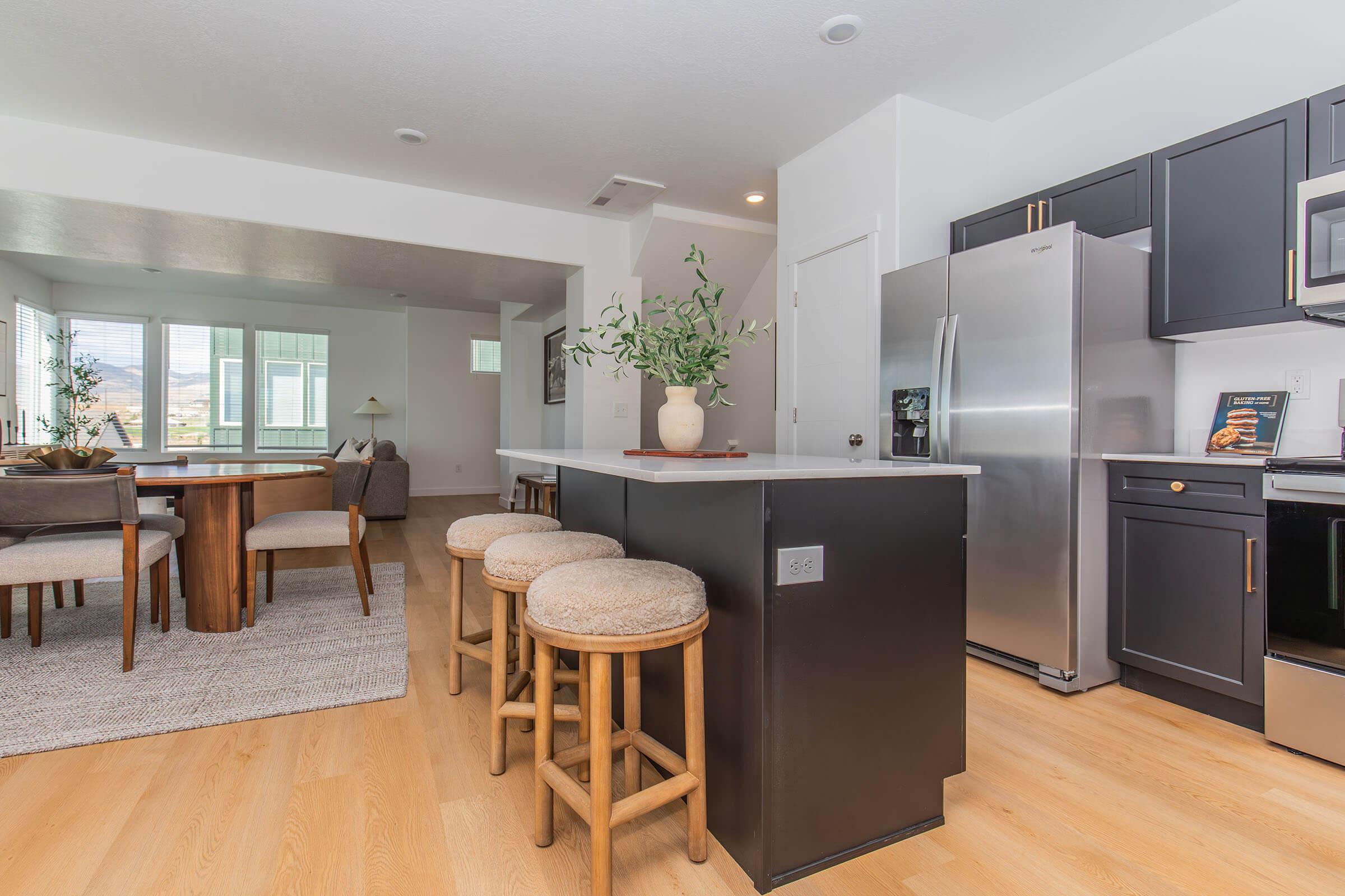 Modern kitchen and dining area featuring sleek black cabinetry, stainless steel appliances, a marble-topped island with three wooden stools, and a dining table set for four. Natural light flows in through large windows, highlighting the open, airy layout and warm wooden flooring.