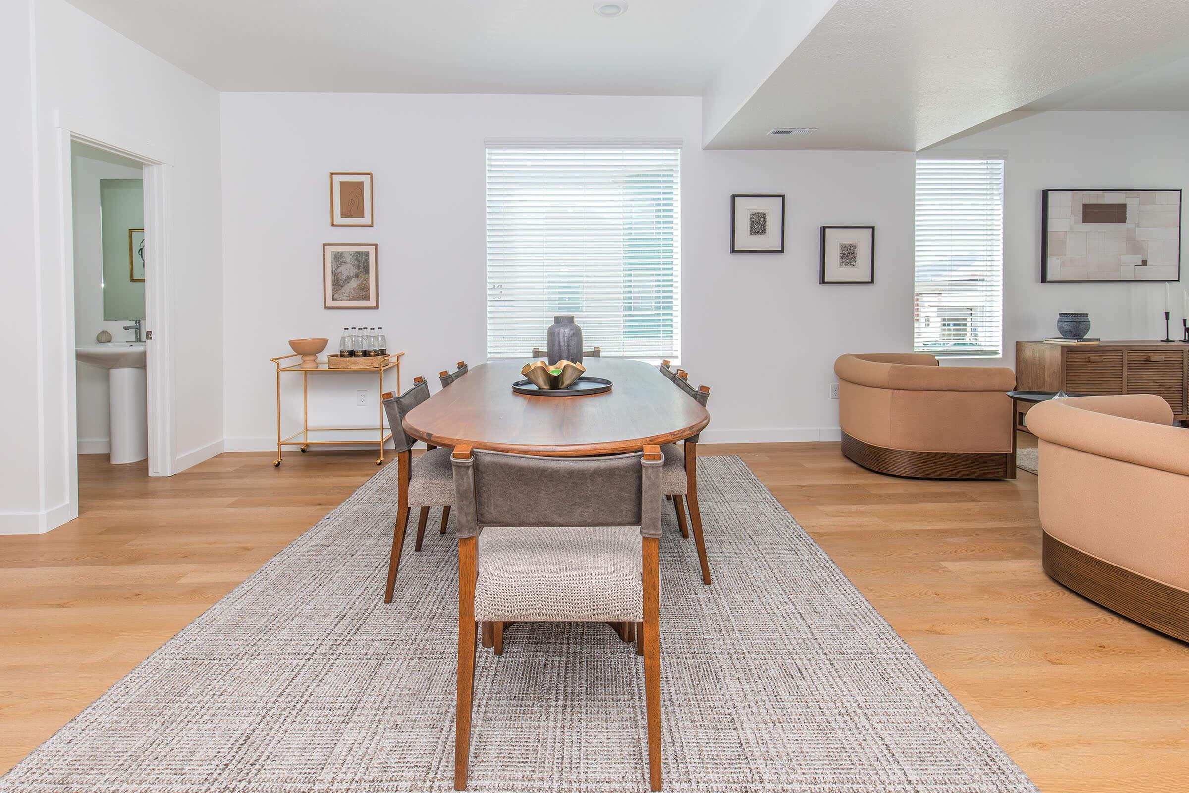 A modern dining room featuring a long wooden table surrounded by upholstered chairs. A decorative bowl sits on the table. Light-colored walls and large windows provide natural light, while framed artworks adorn the walls. A cozy sitting area with curved chairs is visible in the background.