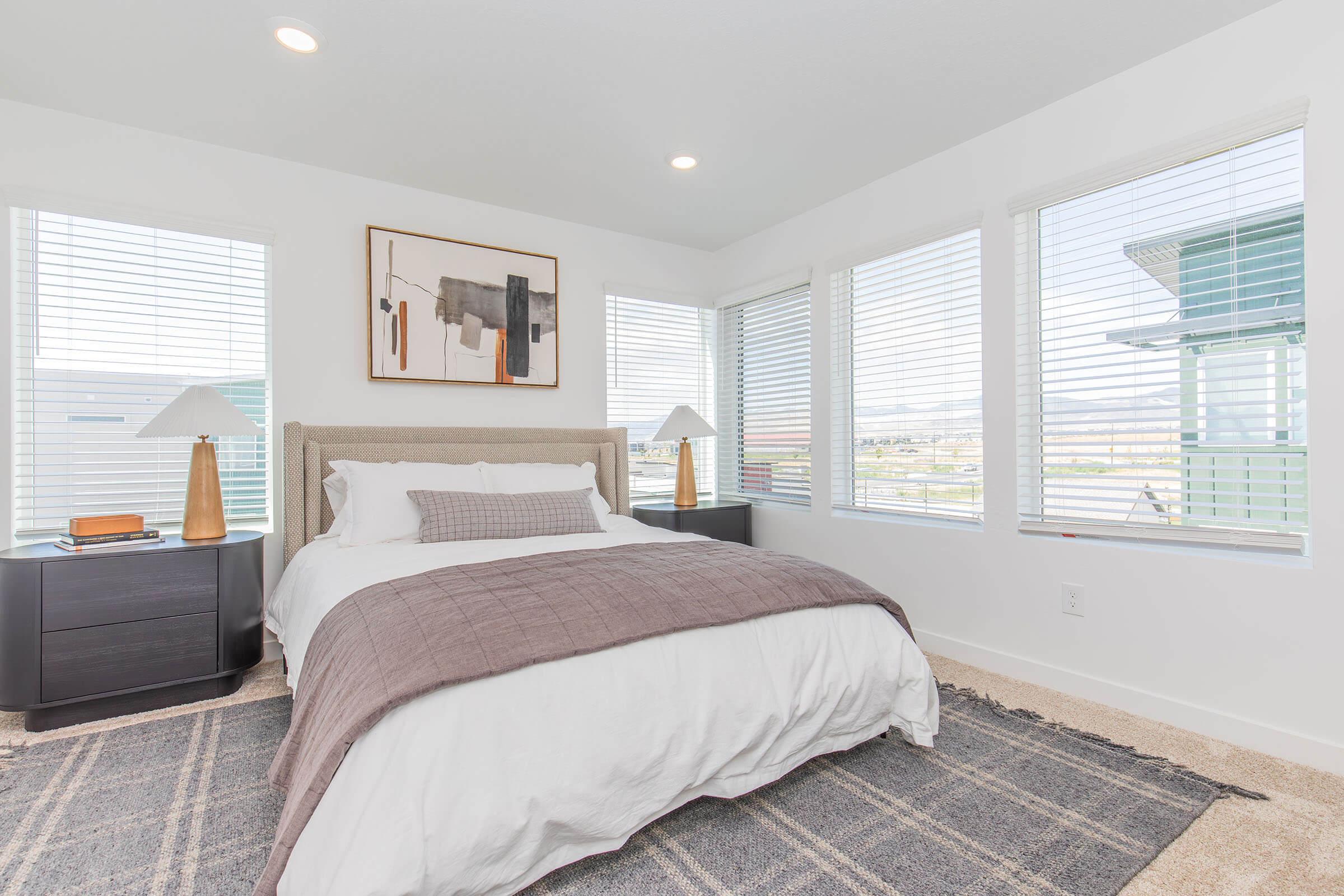 A bright, modern bedroom featuring a queen-sized bed with white bedding and a gray throw blanket. Two table lamps with wooden bases are on either side of the bed, and large windows with white blinds provide natural light. A textured area rug is on the carpeted floor, adding warmth to the space.