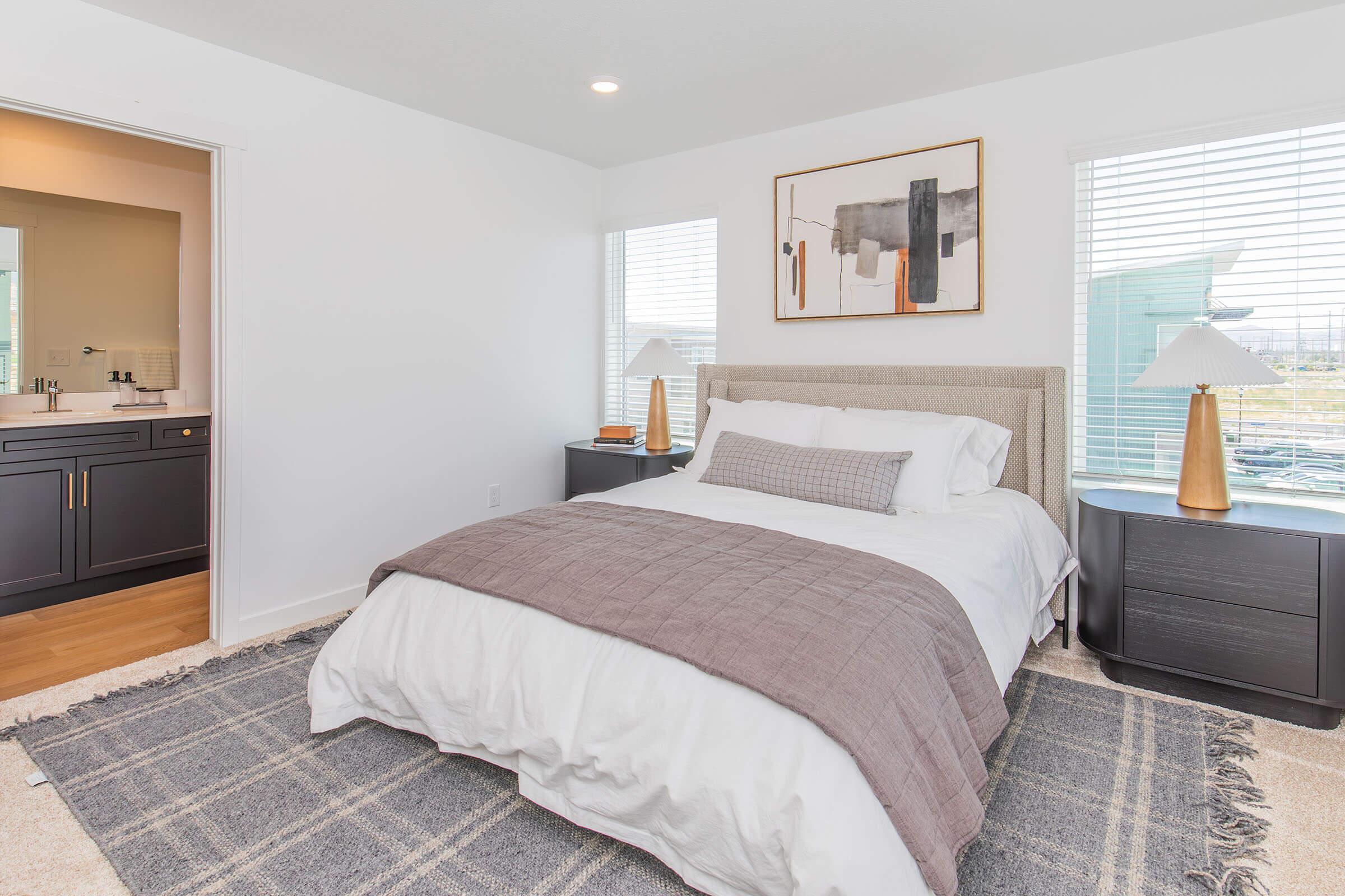 A modern bedroom featuring a queen-sized bed with a light gray blanket, two bedside tables with lamps, and large windows with white blinds. A cozy area rug adds texture to the floor, and an abstract wall art piece is hung above the bed. A bathroom entrance is visible in the background.