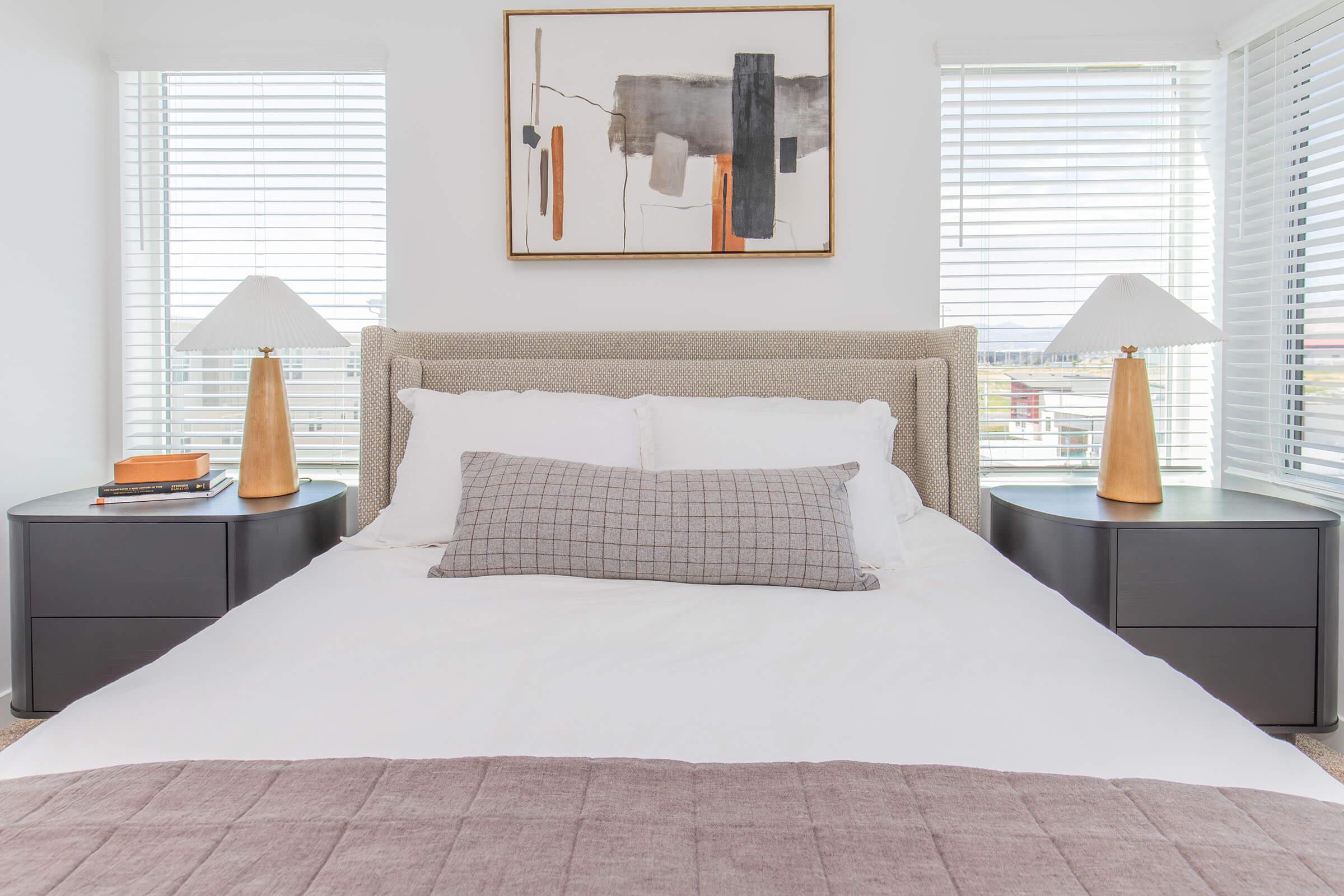 A modern bedroom featuring a neutral color palette. The bed is dressed in white linens with a textured throw blanket at the foot. Two bedside tables with warm-toned lamps are positioned on either side of the bed. A large abstract artwork hangs above the headboard, and sunlight streams through the window blinds.