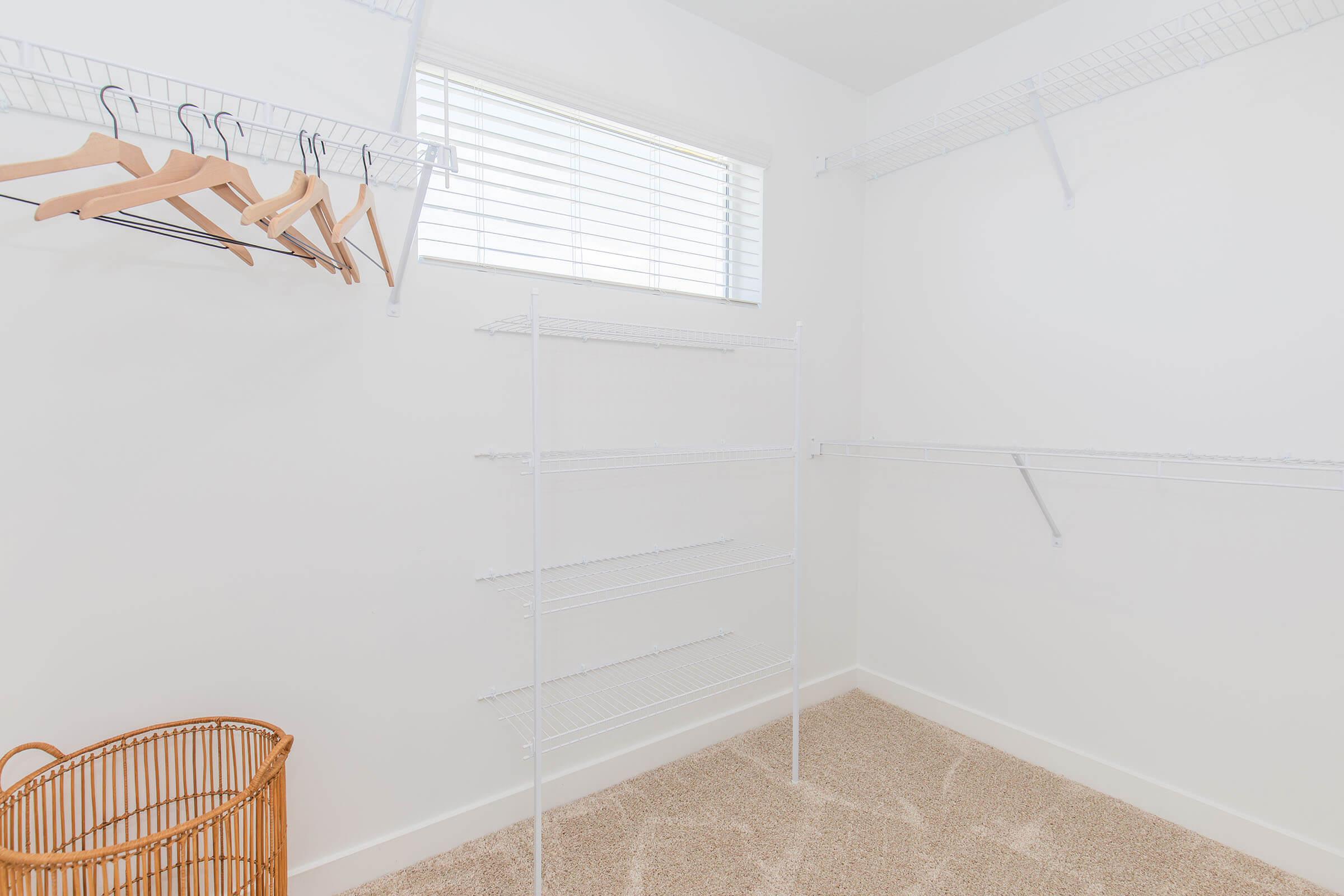 A clean and empty closet showcasing white wire shelving, a few wooden hangers hanging from the wall, a large woven laundry basket, and a small window allowing natural light to filter in. The walls are painted white, and the floor is covered in beige carpet.