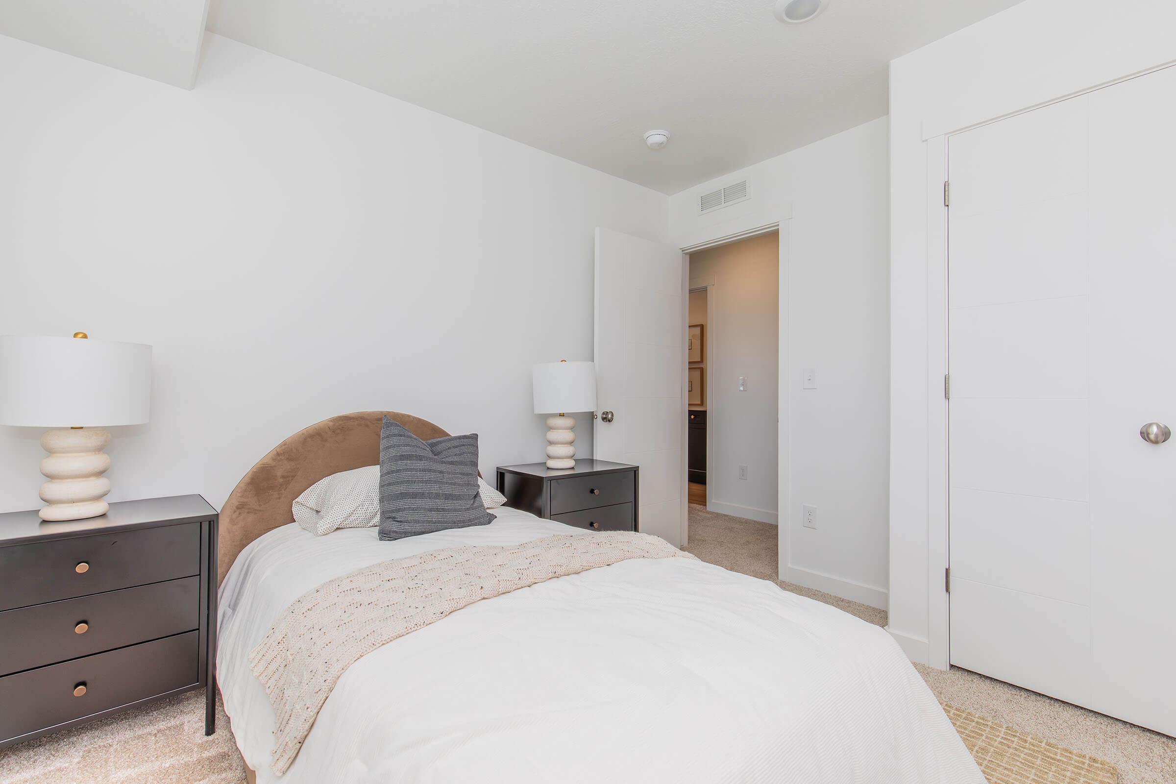 A neatly designed bedroom featuring a single bed with white bedding, adorned with a beige throw. Two stylish nightstands with lamps are on either side of the bed. The walls are painted white, and there is a closed door leading to another room, adding to the airy and modern feel of the space.