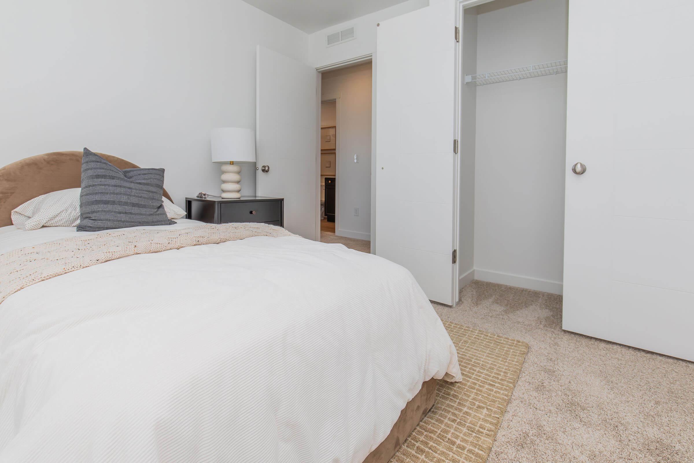 A cozy bedroom featuring a queen-sized bed with white linens and a decorative throw pillow. A bedside table with a lamp is next to the bed, and a spacious closet stands open with sliding doors. The room has neutral-colored walls and carpet, creating a warm and inviting atmosphere.