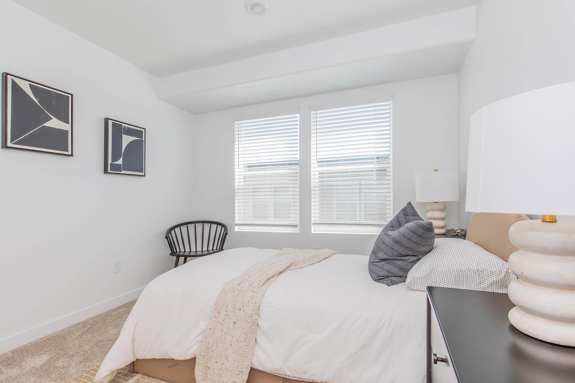 A modern bedroom featuring a neutral color palette, a comfortable bed with decorative pillows, a nightstand with a lamp, and a stylish chair. Large windows with white blinds let in natural light, while framed artwork adds a touch of elegance to the space. A cozy throw is draped over the bed.