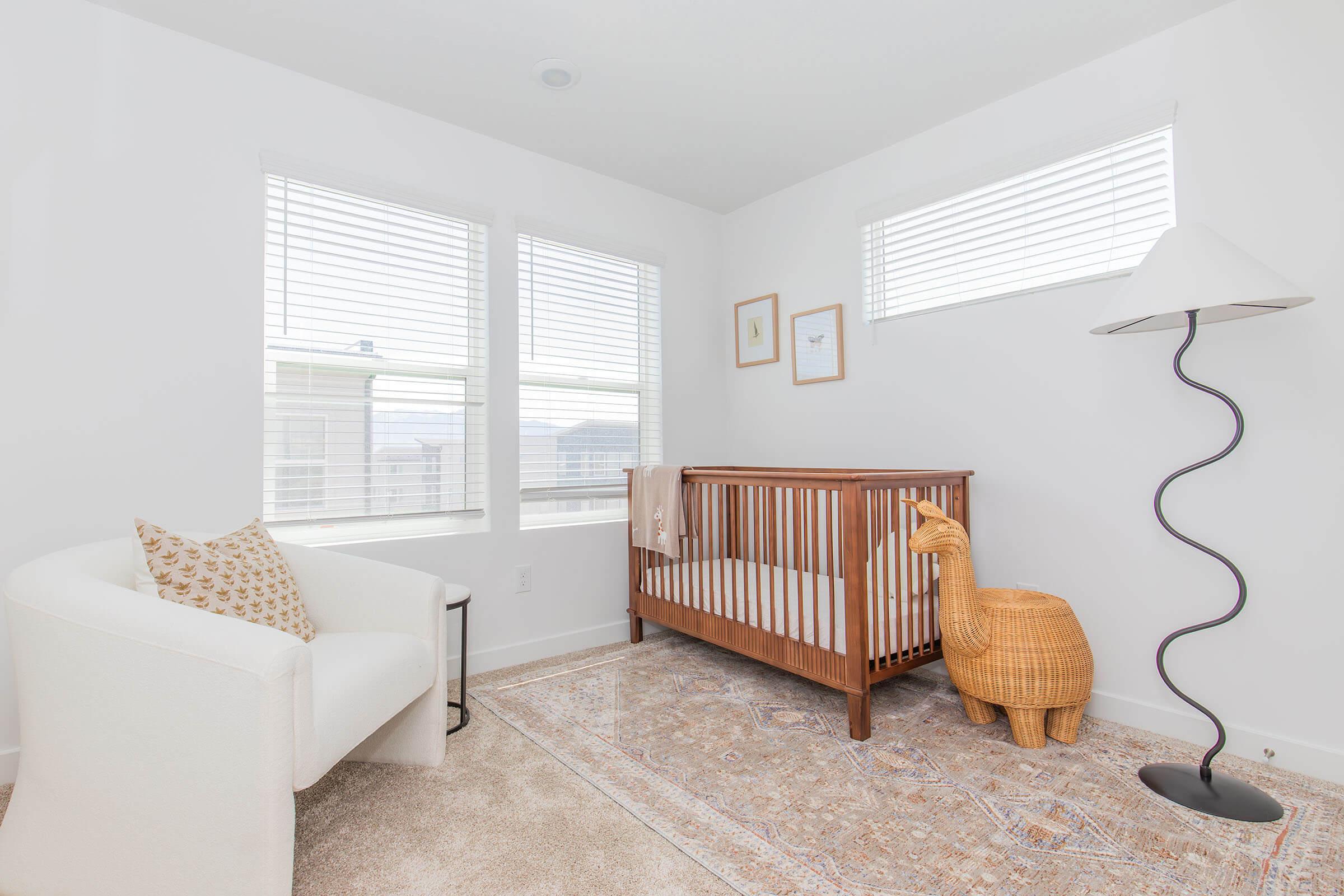 A bright, modern nursery featuring a wooden crib, a unique wicker animal-shaped decor, a comfortable white armchair, and a stylish floor lamp. Large windows allow natural light to fill the room, and a patterned rug adds warmth to the space. Soft, neutral color palette enhances the calming atmosphere.