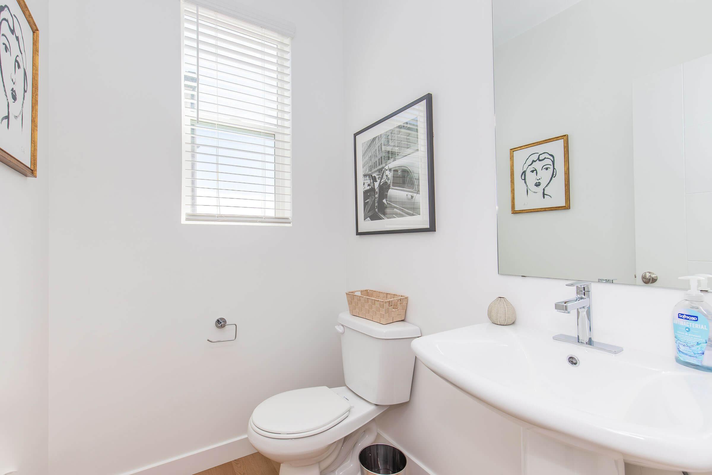 A modern bathroom featuring a white toilet, a sink with a mirror, and a small waste basket. A window with blinds allows natural light to enter, and there are two framed artworks on the wall, including a minimalist drawing and a black-and-white photograph. The decor is clean and contemporary.