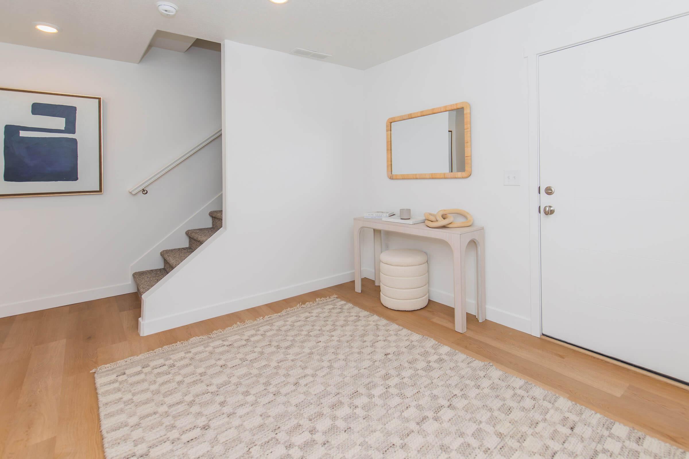 A modern entryway featuring a white wall, a simple console table, a round stool, and a decorative mirror. A staircase leads to the upper level, and an abstract painting is hung on the wall. A textured area rug adds warmth to the space against the light wooden flooring.