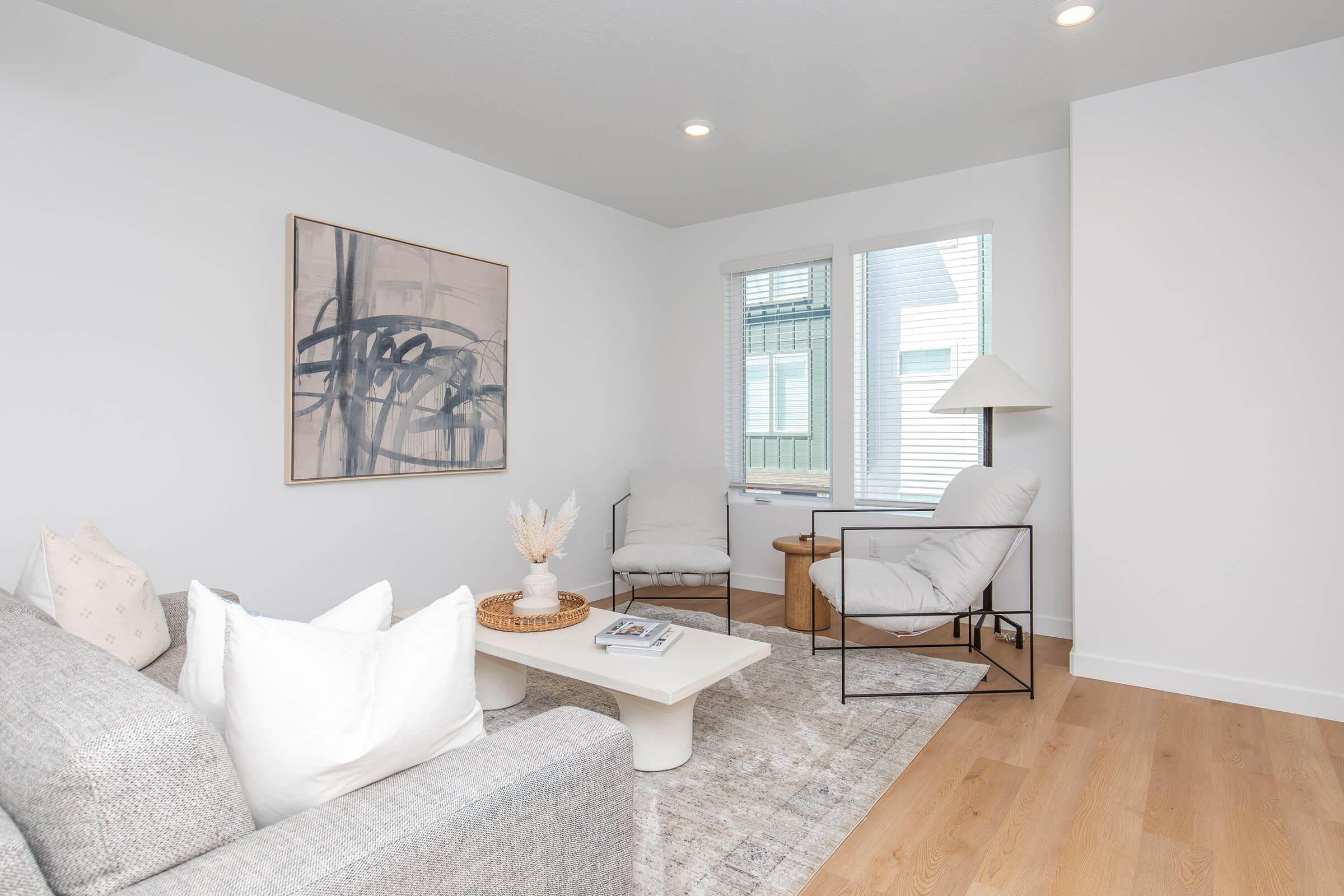 A modern and airy living room featuring a light gray sofa with decorative pillows, a white coffee table, and two stylish chairs. Natural light streams in through large windows, illuminating the minimalist decor, which includes a piece of abstract art on the wall and a small plant.