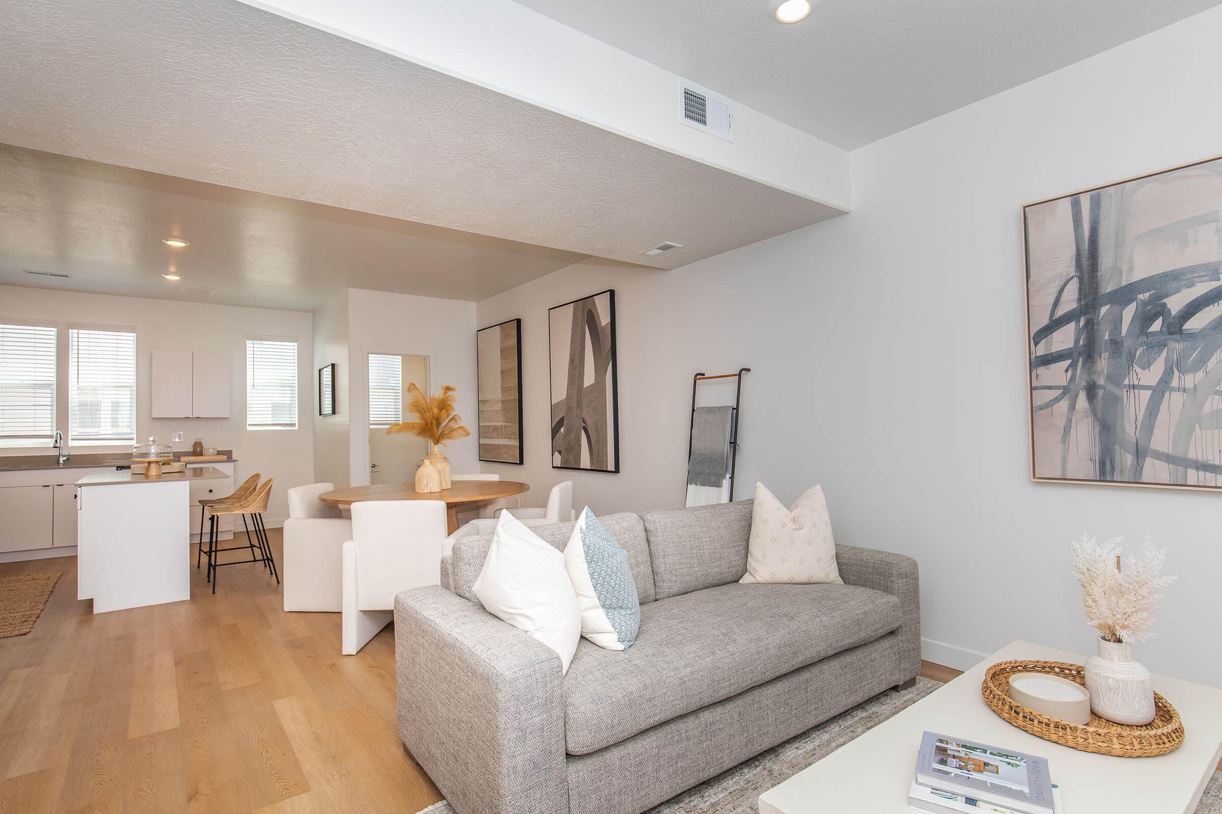 A modern living area featuring a light gray sofa adorned with decorative pillows. In the background, an open kitchen with white cabinetry is visible, alongside a dining area with a round table and chairs. Natural light streams through large windows, enhancing the clean, minimalist decor.