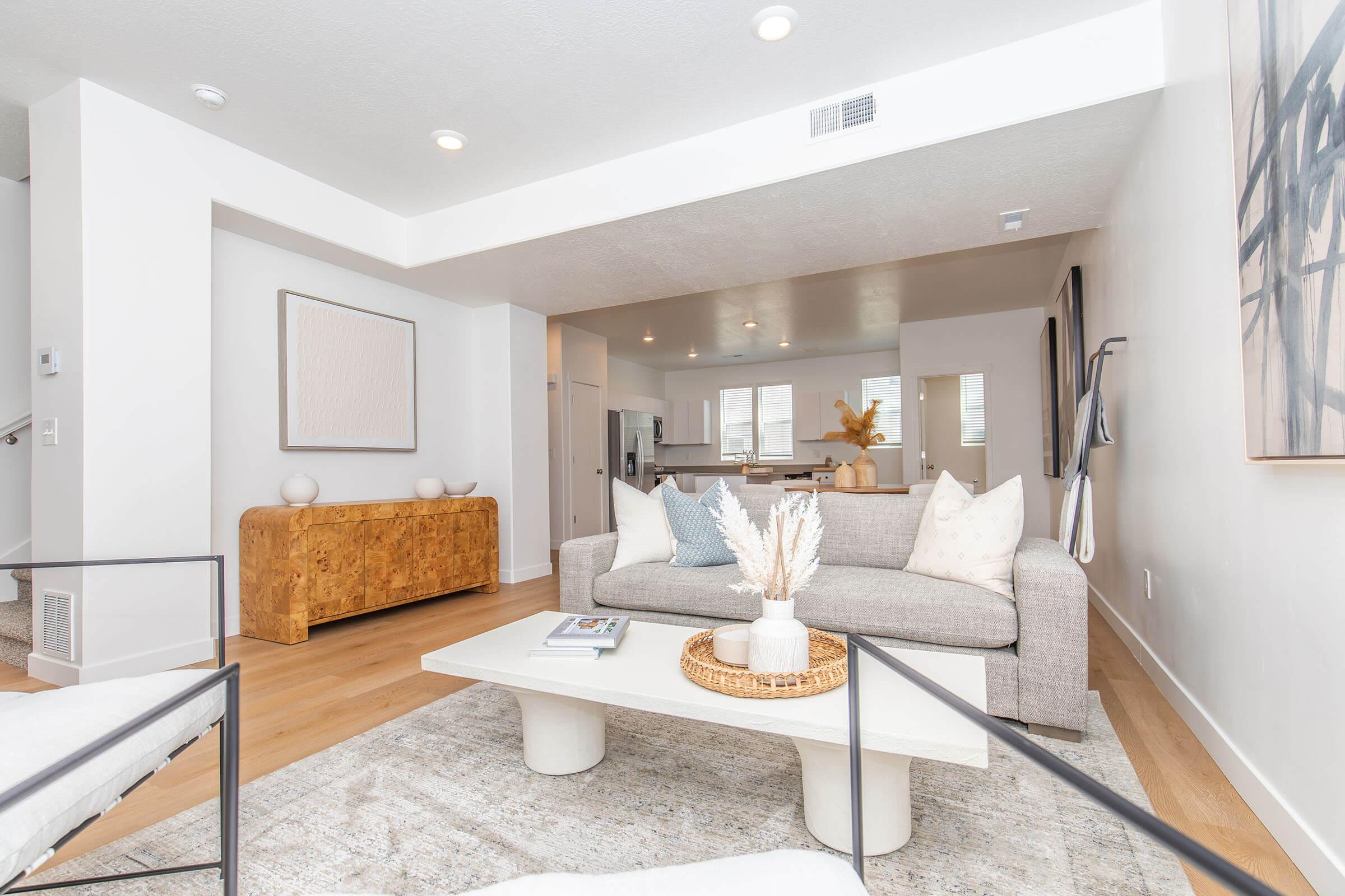 Modern living room featuring a light gray sofa with decorative pillows, a white coffee table on a textured rug, and a wooden sideboard. Natural light streams in from windows, highlighting minimalist decor and a cozy atmosphere. The space is designed with neutral tones and contemporary furnishings.