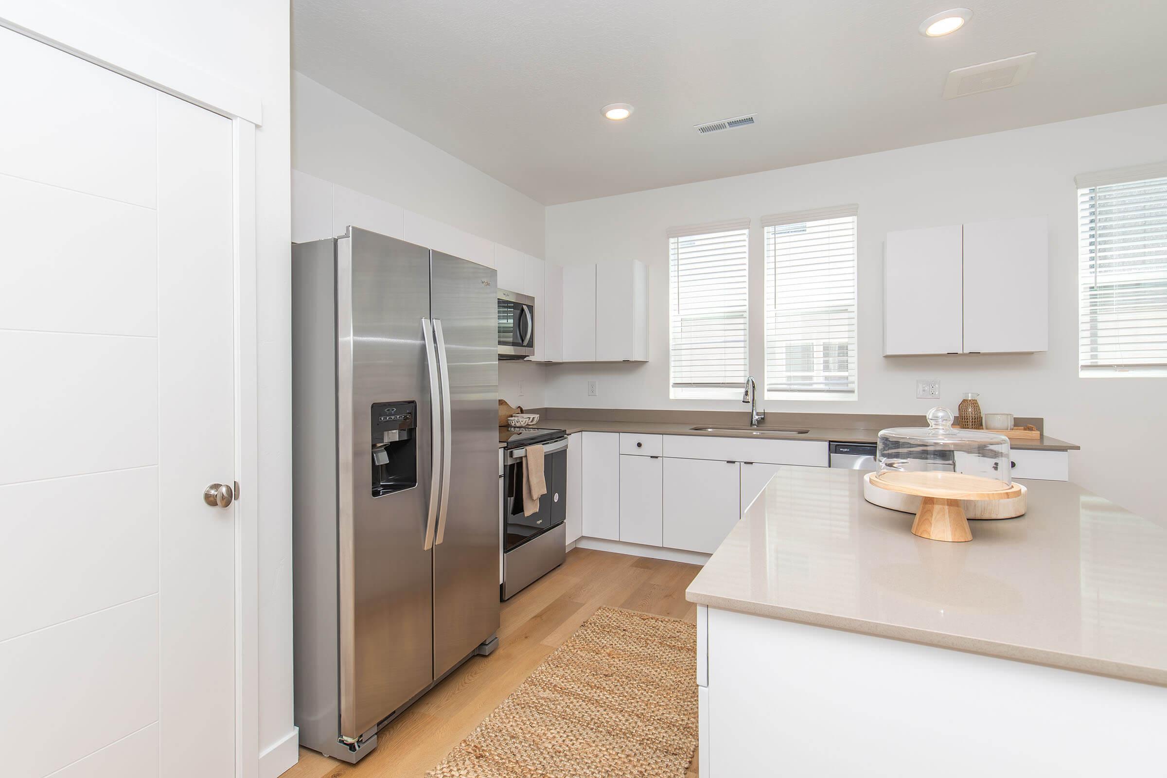 A modern kitchen featuring stainless steel appliances, including a refrigerator and oven. The cabinetry is white with a clean, minimalist design, and large windows allow natural light to illuminate the space. A wooden countertop and a woven rug add warmth to the contemporary look.