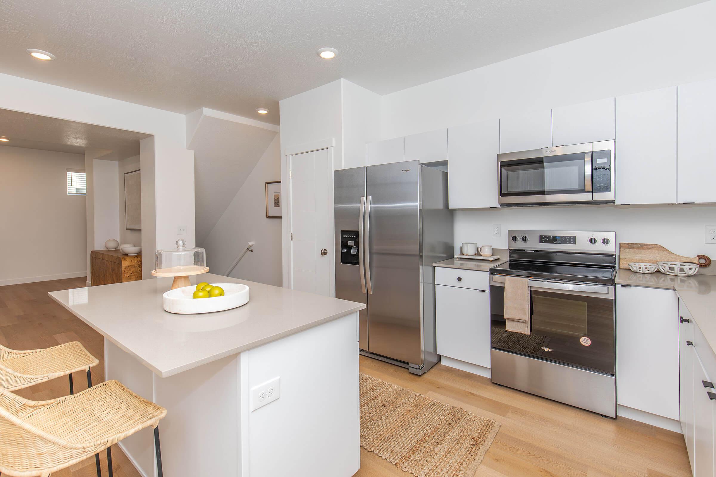 Modern kitchen featuring stainless steel appliances, including a refrigerator, oven, and microwave. The space has white cabinetry, a light countertop, and a small island with three wicker bar stools. A neutral rug lies on the wooden floor, and decorative items are placed throughout the area, contributing to a clean and contemporary look.