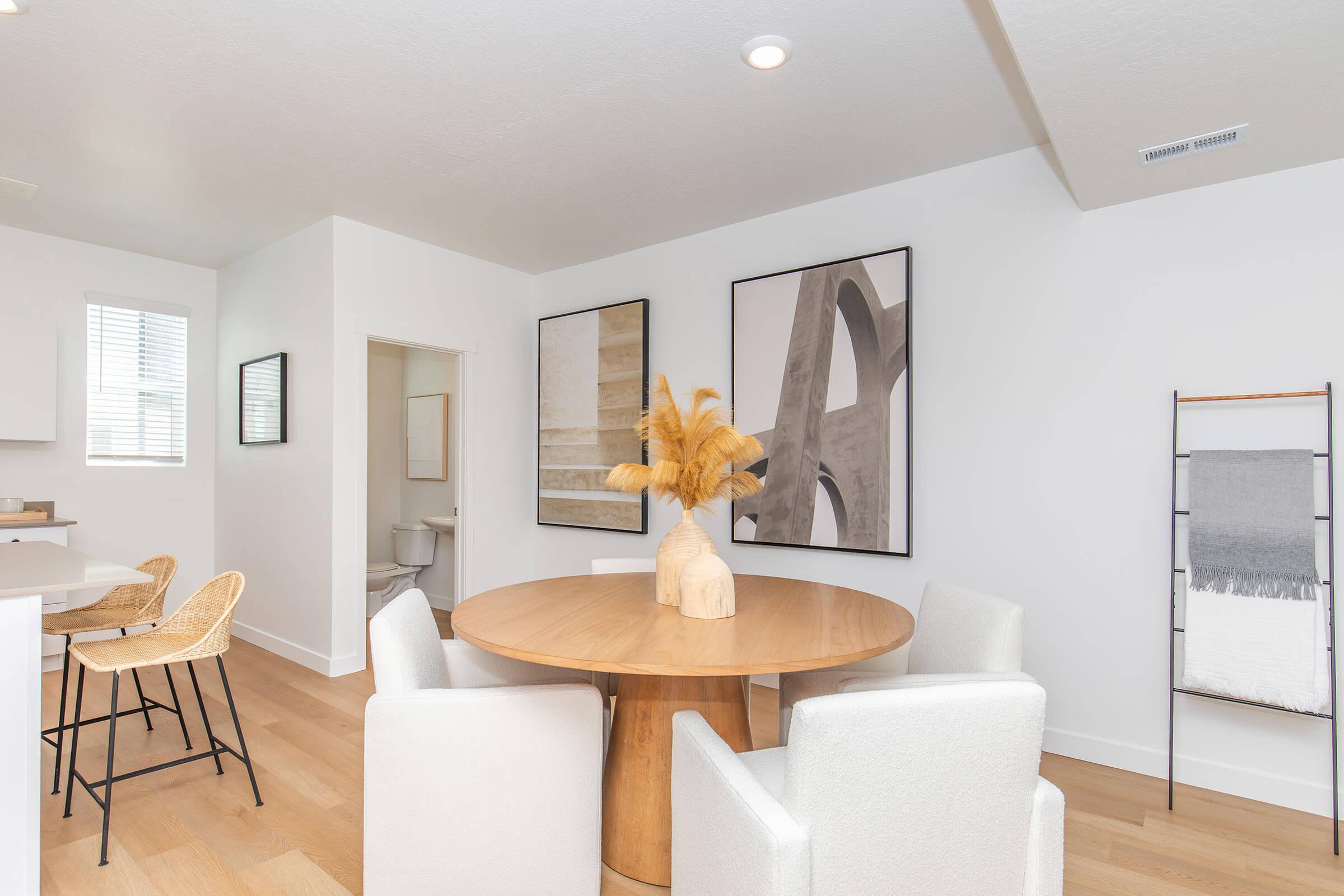 Contemporary living space featuring a round wooden dining table surrounded by white chairs. A modern vase with decorative dried flowers sits at the center. The walls are adorned with abstract art pieces, and there's a glimpse of a bathroom in the background. Bright, minimalistic design with light wood flooring.