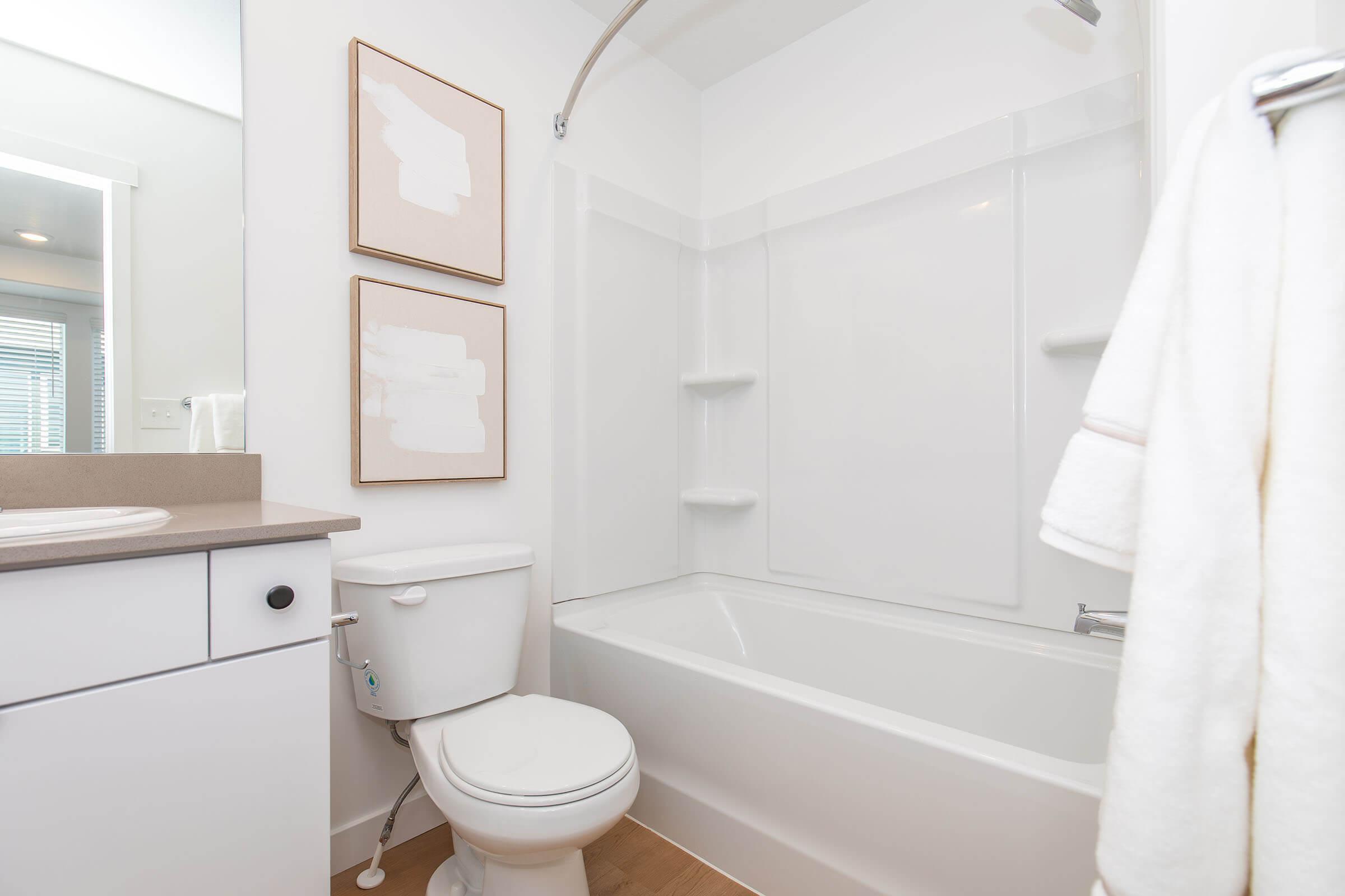 A modern bathroom featuring a bathtub with a shower, a toilet, and a sink with a countertop. Two abstract art pieces are mounted on the wall. Soft lighting enhances the clean, contemporary aesthetic, with light-colored walls and wooden accents on the floor. A neatly hung bath towel is visible.
