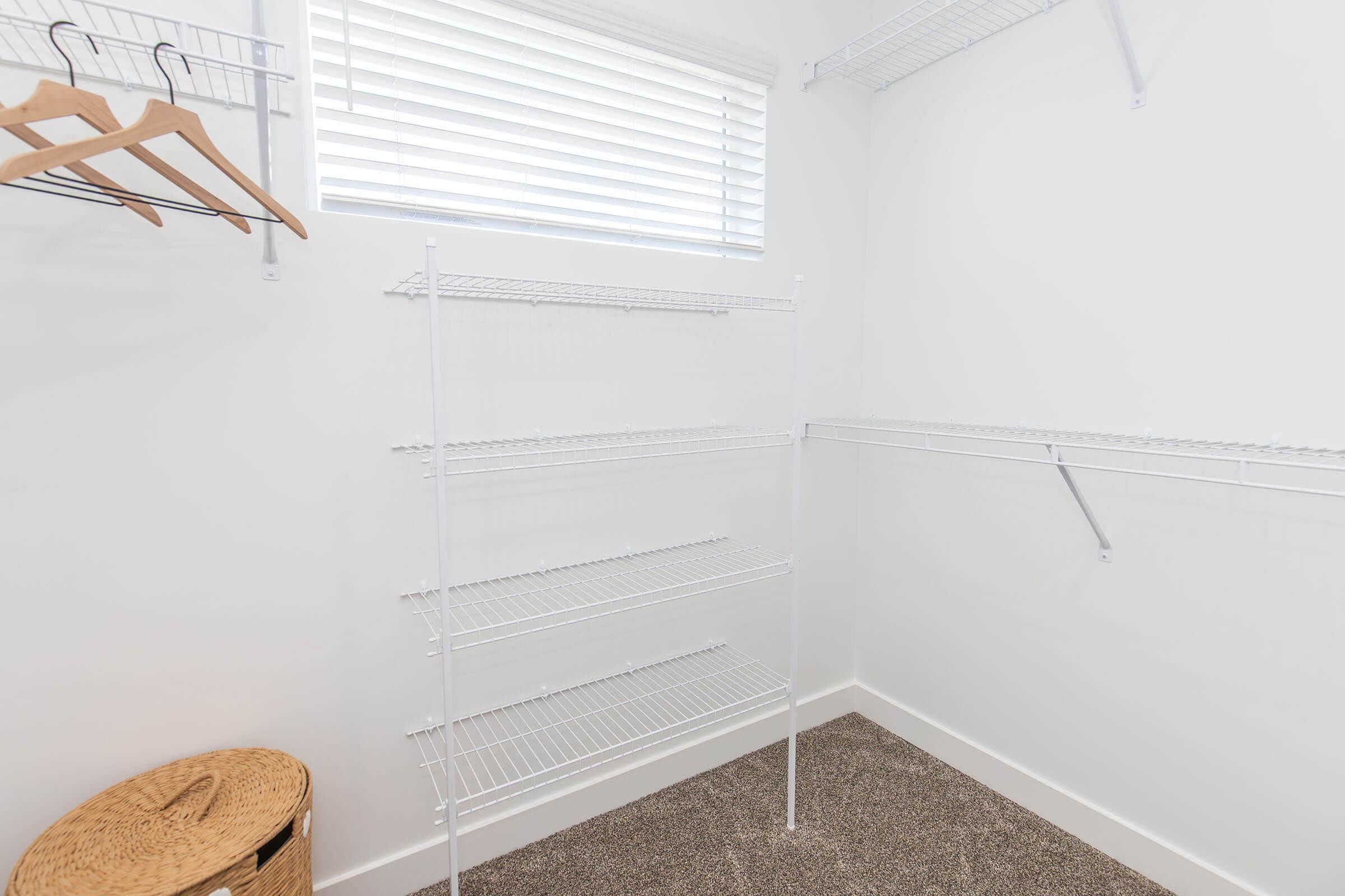 A clean, empty closet with white wire shelving units and a wicker basket on the floor. There are wooden hangers hung on the rail, and a window with white blinds provides natural light. The walls are painted white, and the floor is covered with light-colored carpet.