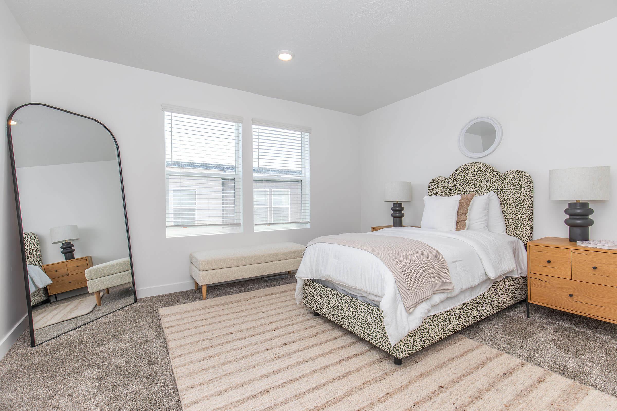 A bright and modern bedroom featuring a patterned bed with white bedding, two bedside lamps, and a large mirror. There is a light-colored bench at the foot of the bed and a soft area rug on the carpeted floor. Two windows allow natural light to fill the space.