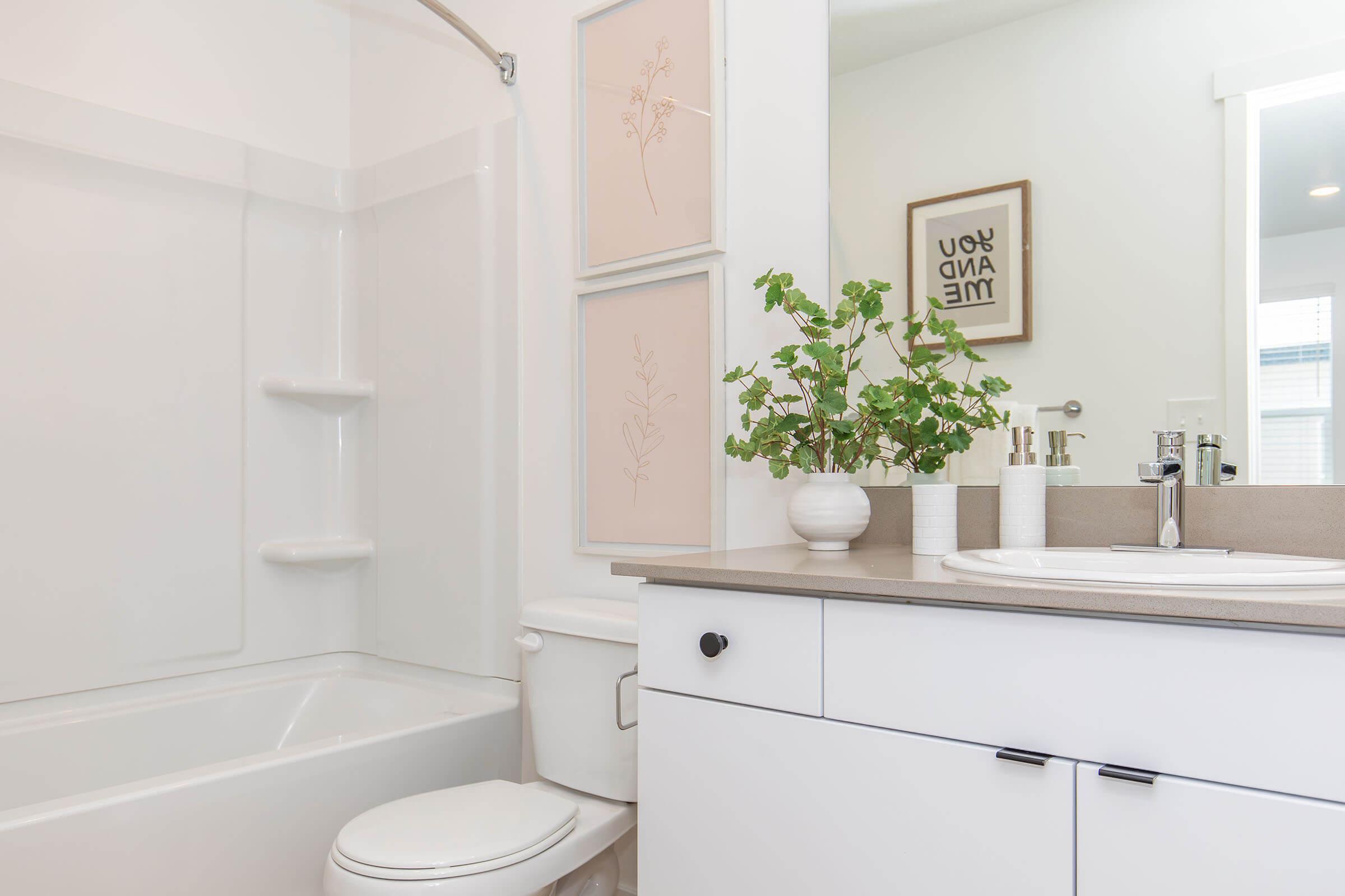 A modern bathroom featuring a white bathtub with a shower, a sink with a large mirror, and minimalistic decor. The counter has potted greenery and decorative vases. Light-colored walls and natural light from a window create a bright, airy atmosphere. Framed art is displayed on the walls.