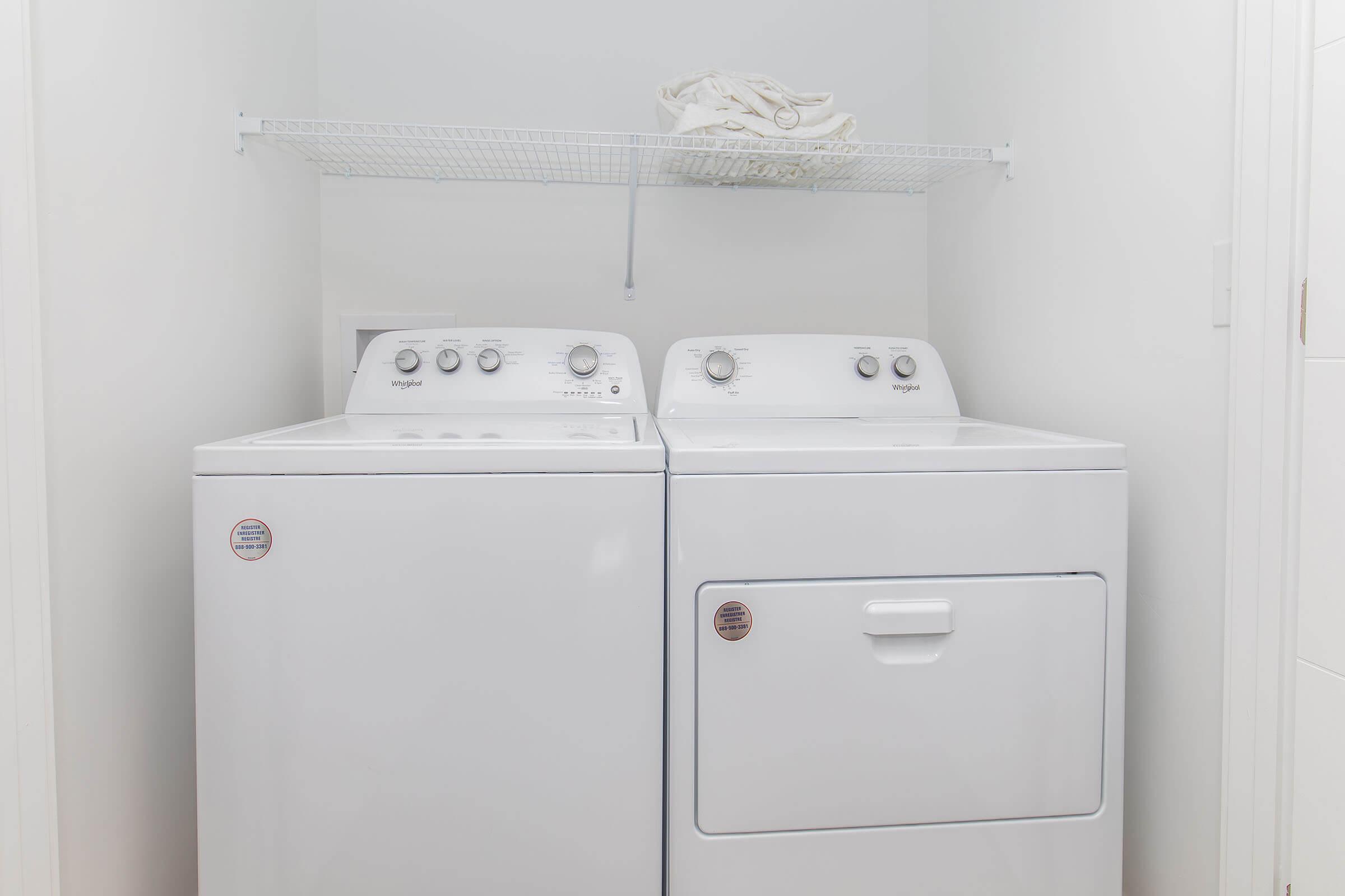 A white washing machine and dryer side by side in a laundry room. Above them is a wire shelf holding folded towels. The walls are painted white, creating a clean and modern look.