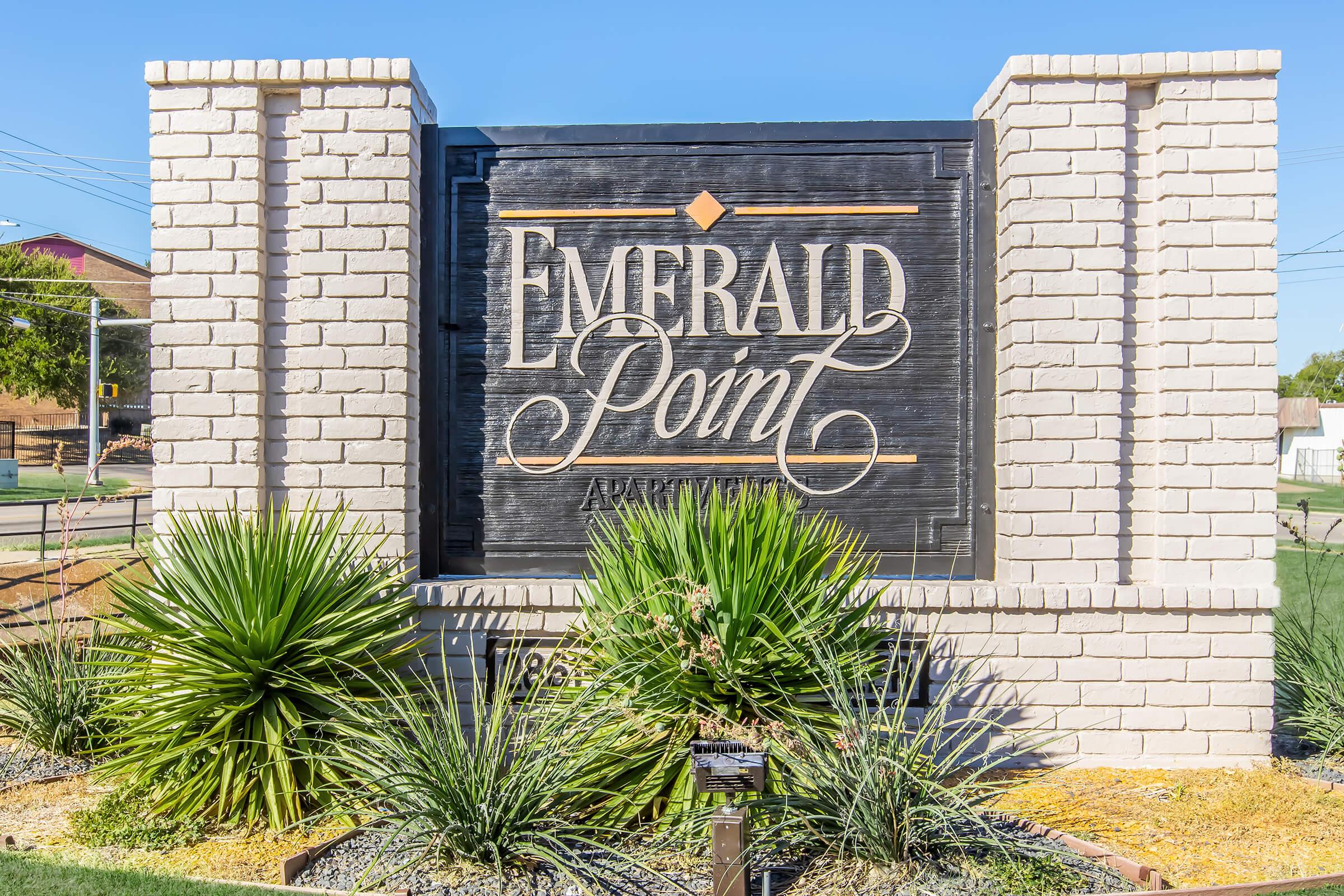 Sign for "Emerald Point" with elegant typography and a decorative diamond design. Surrounded by various types of landscaping, including green shrubs and spiky plants. The background is clear blue sky, indicating a sunny day.