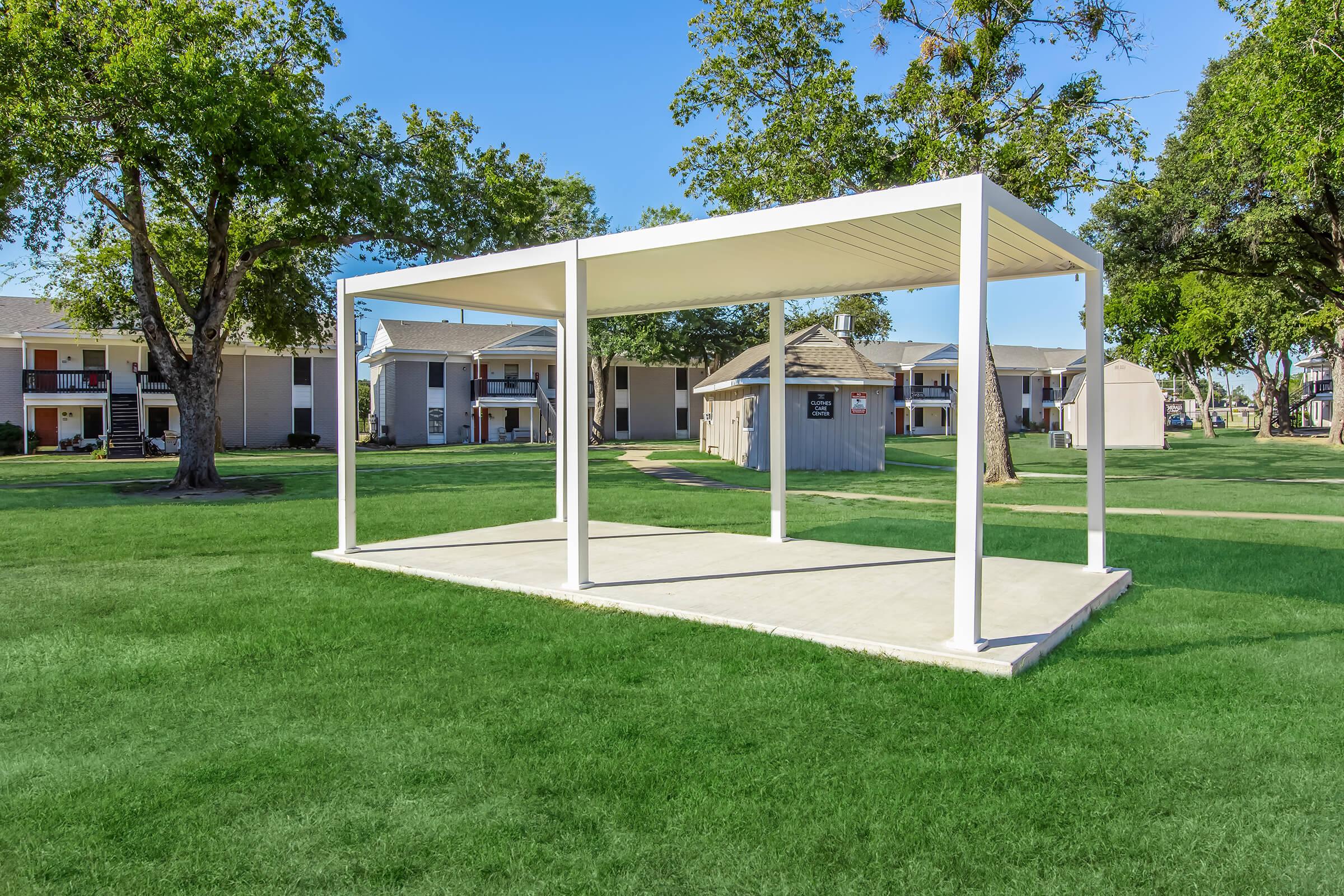 A spacious outdoor structure with a white frame and a concrete base, situated on a grassy area. Nearby, there are residential buildings with a few trees providing shade, creating a serene environment. The sky is clear and blue, enhancing the inviting atmosphere of the space.
