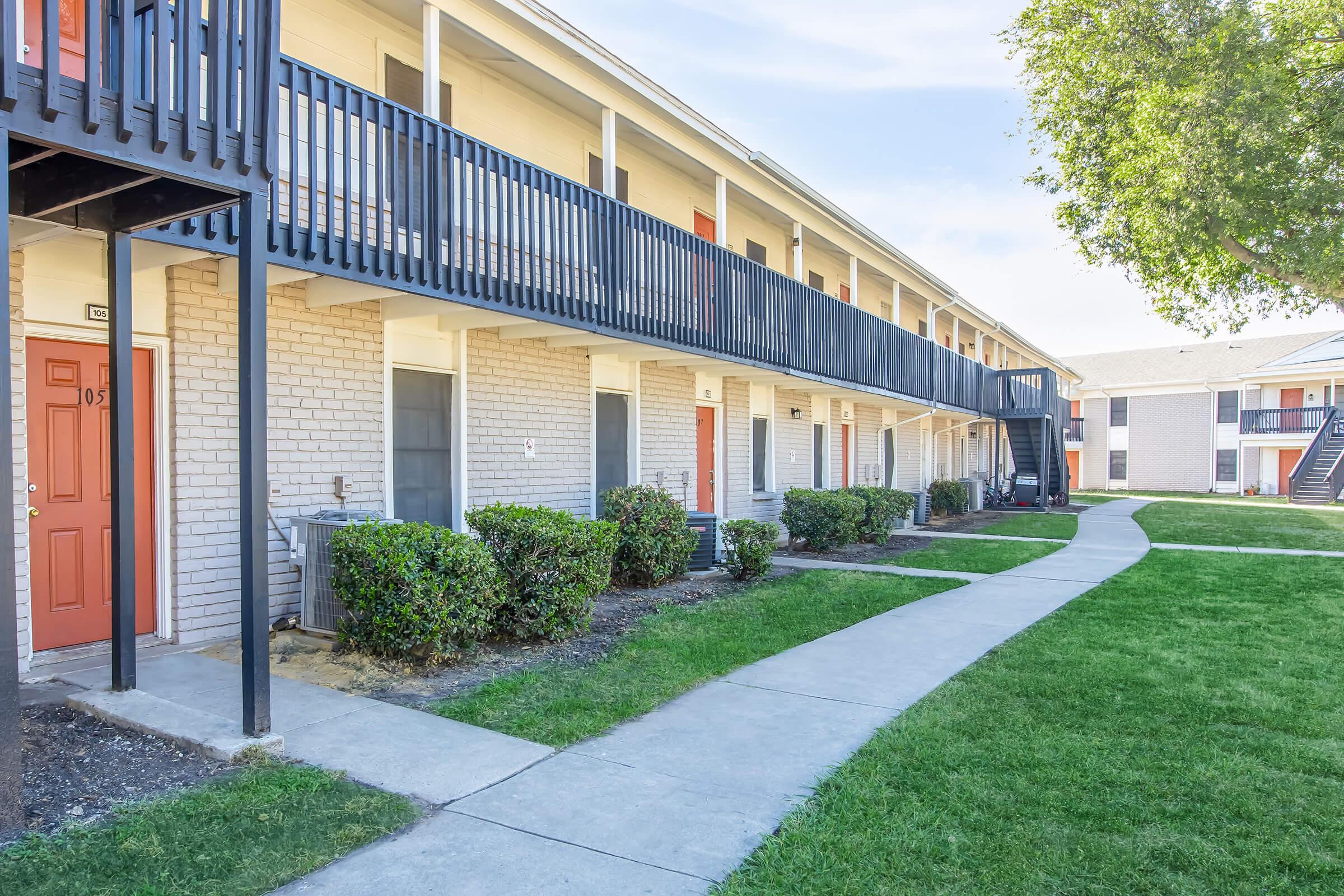 A row of apartments featuring exterior balconies and red doors, with a paved walkway leading through well-maintained grass and shrubs. The building is two stories high, and there is a staircase on the right leading to the upper level. The setting appears clean and inviting.