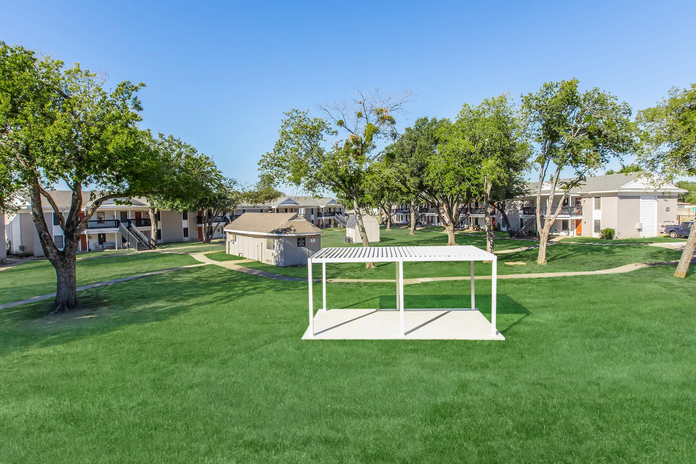 A spacious green lawn surrounded by apartment buildings, featuring several trees. In the foreground, there is a white gazebo-style structure on a concrete slab, providing a shaded area. The clear blue sky adds to the serene atmosphere of the setting.