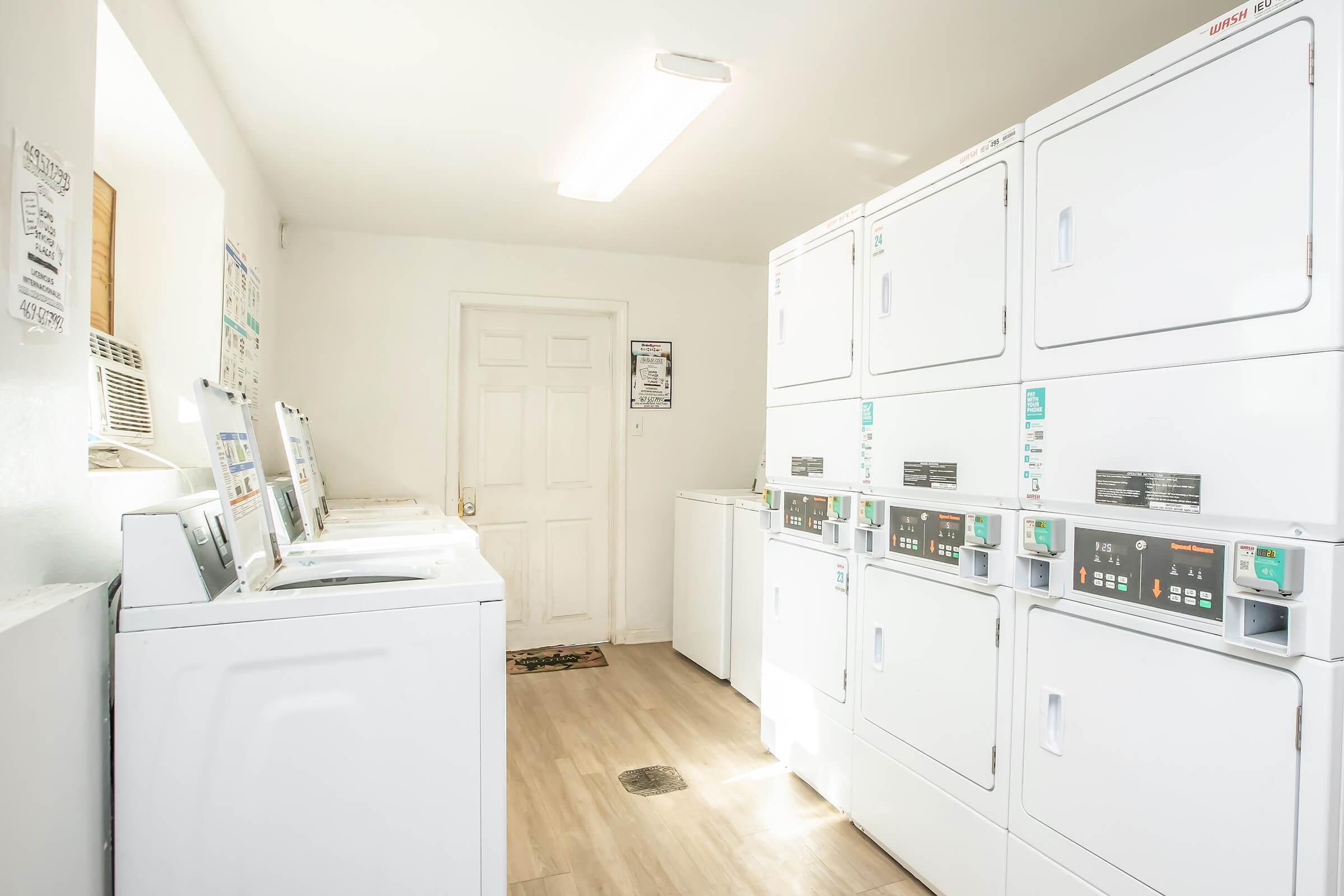A bright, clean laundry room featuring several white washing machines and dryers along one wall. There is a door at the end of the room, and an air conditioning unit is visible. The floor is wooden, and the space is well-lit with overhead lighting.