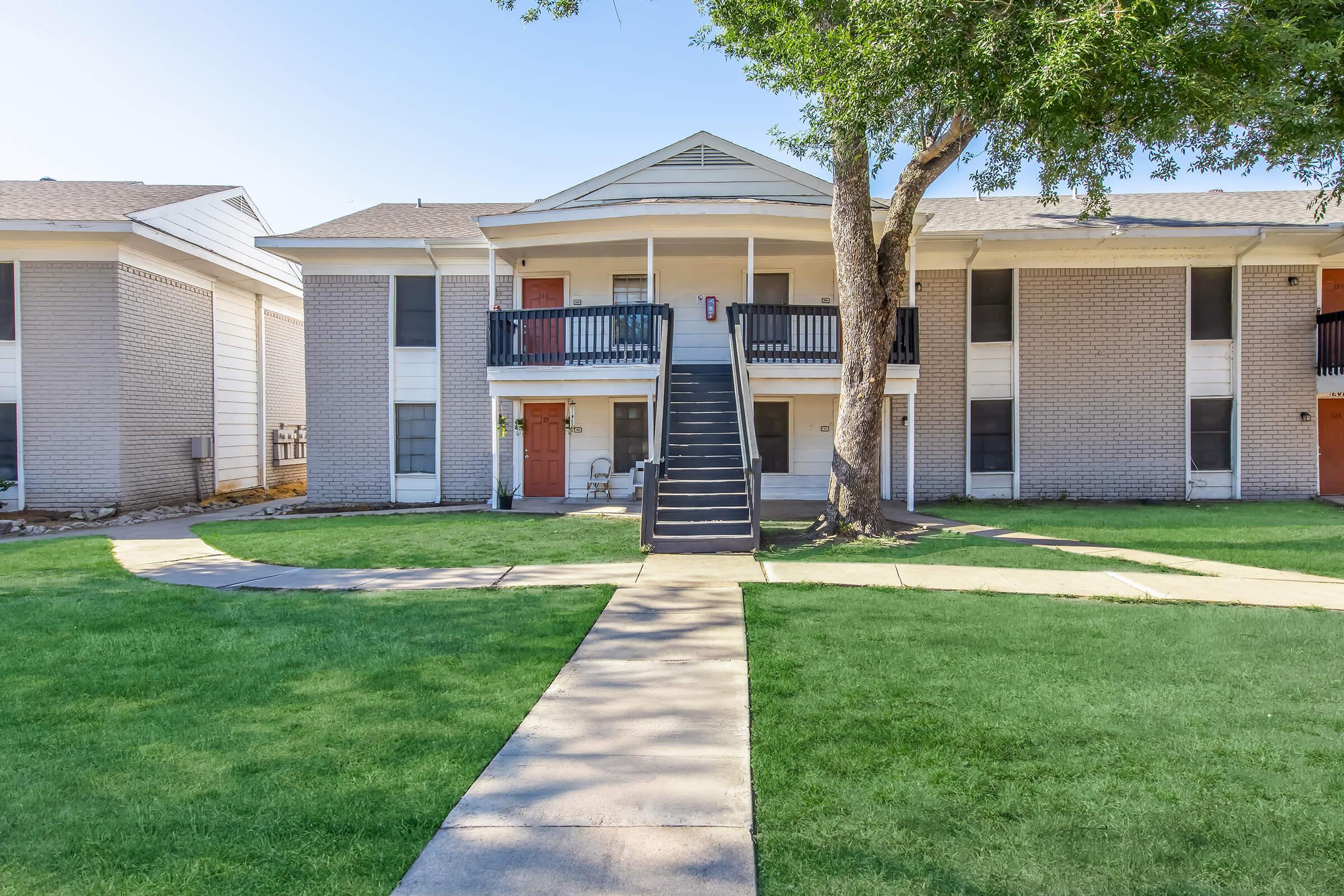 Two-story apartment building with a central staircase leading to the upper floor. The building features a light-colored brick exterior with black railings and doors, surrounded by well-maintained green grass and concrete pathways. There are trees providing shade in the area.