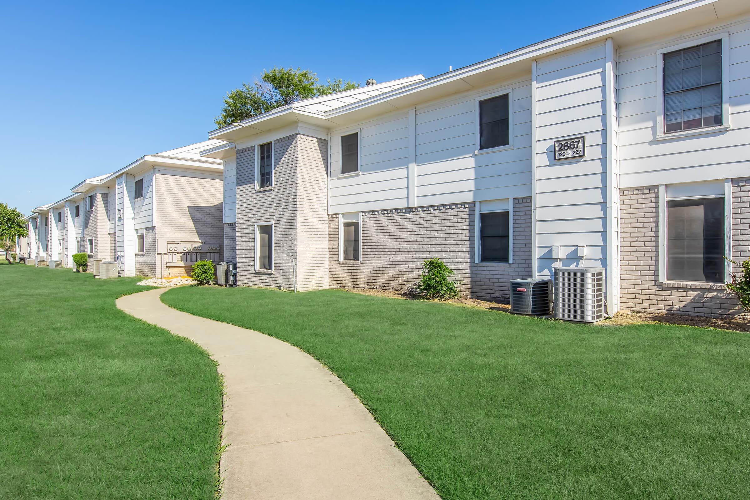 A well-maintained apartment complex with a sidewalk winding through a green lawn. The buildings are two stories with light-colored exteriors, windows, and air conditioning units. Clear blue sky overhead enhances the welcoming appearance of the property.