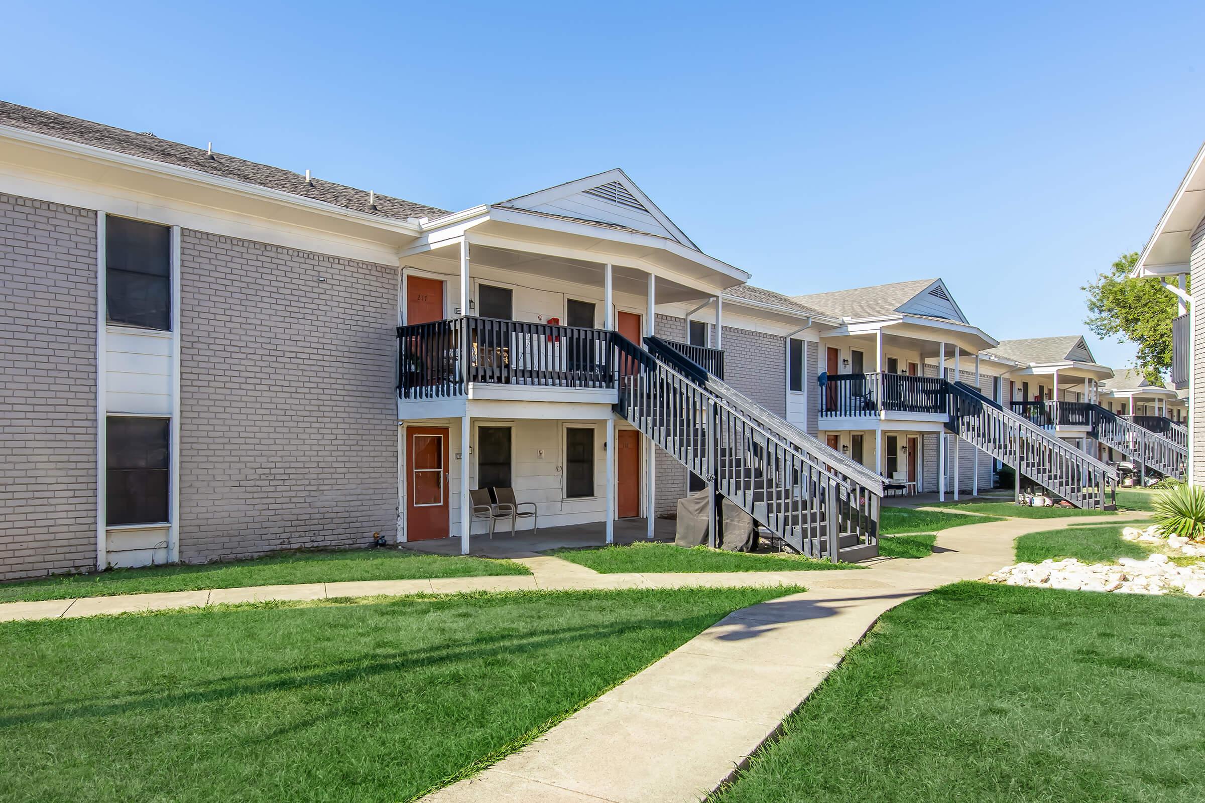 Apartment complex with multiple units featuring elevated entrances, black wooden staircases, and brick exteriors. Lush green lawn and walkways connect the buildings under a clear blue sky.