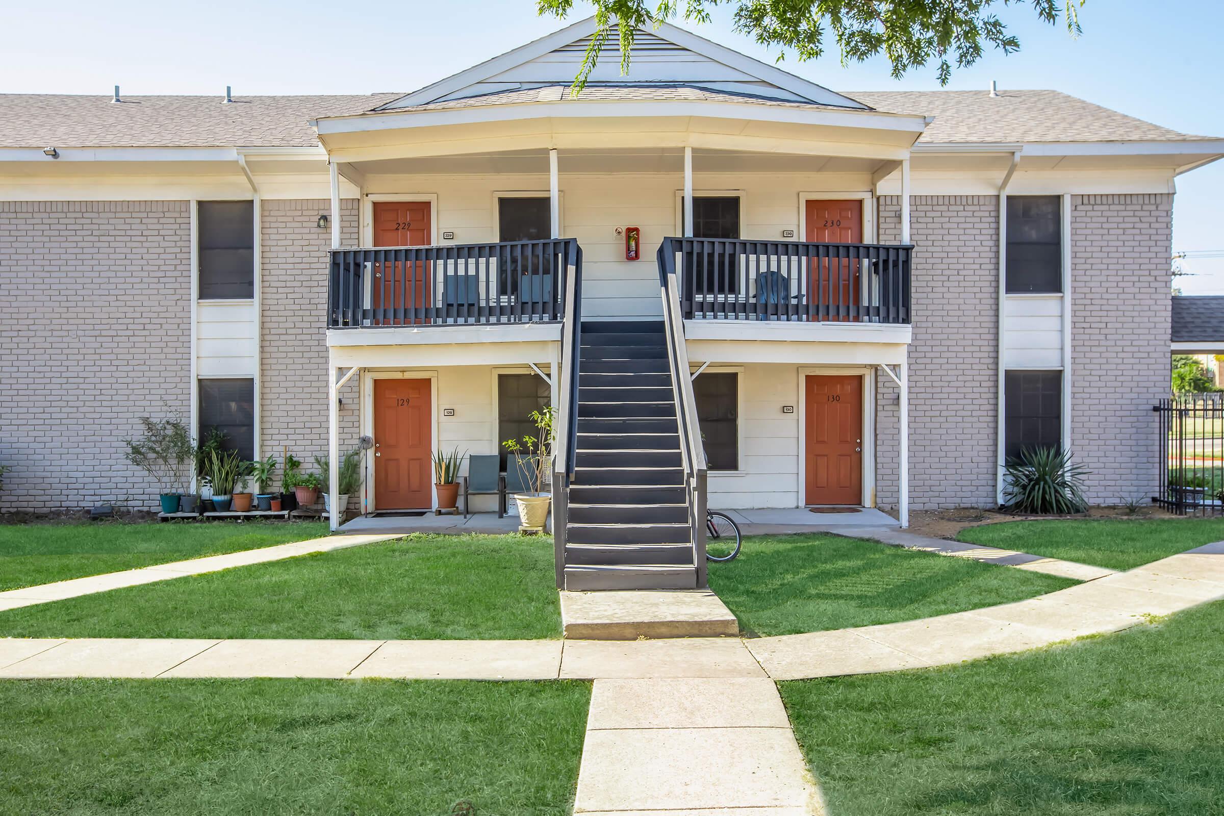 Two-story apartment building with a light gray brick exterior. There are two sets of wooden doors painted orange, leading to individual units. A staircase in the center leads to the upper level. The surrounding area has a well-maintained lawn and some potted plants, with a pathway curving around the building.