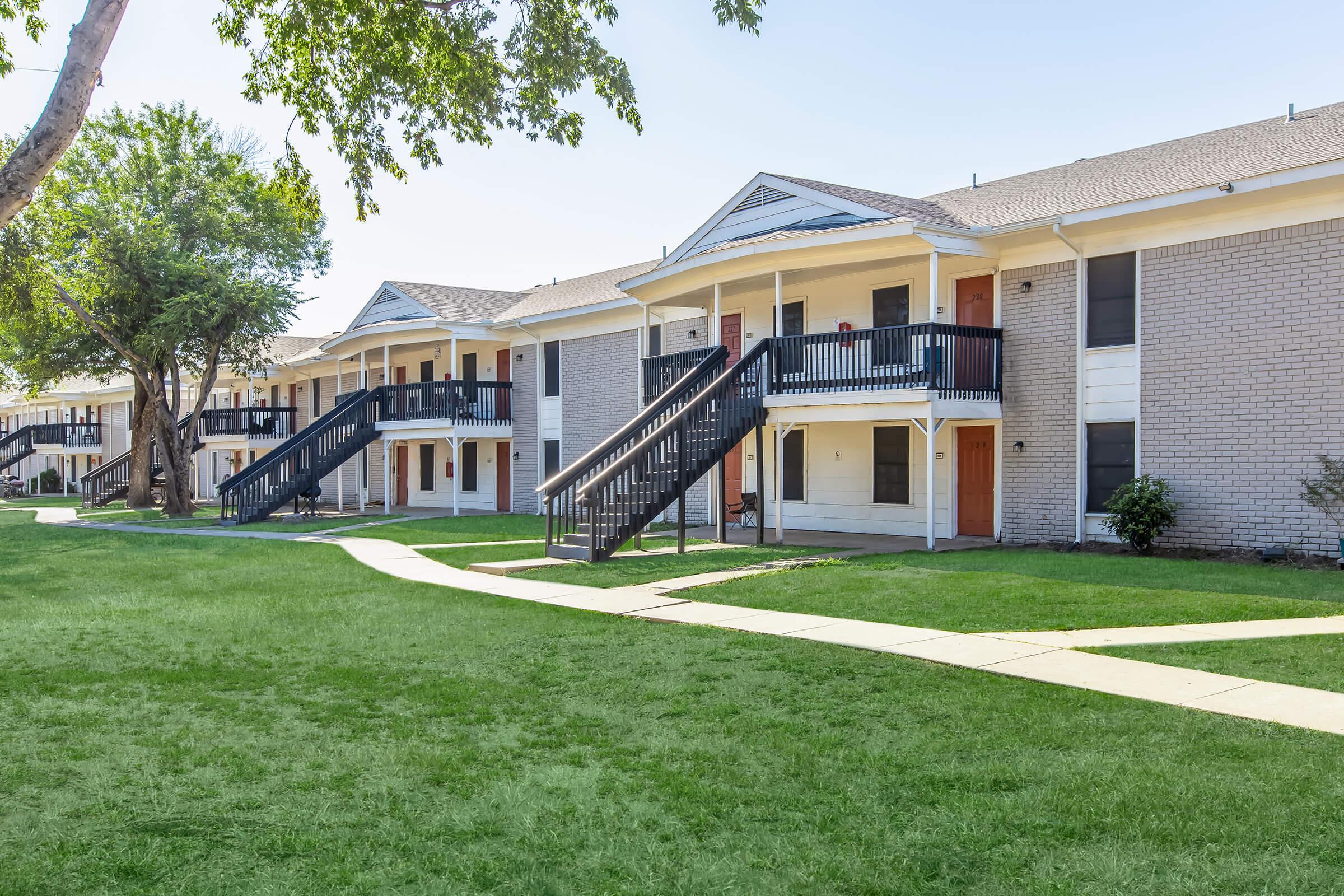 A row of apartment buildings with covered balconies and black railings, surrounded by well-maintained green grass. A concrete pathway winds between the buildings, and trees provide shade in the area. The architecture features a mix of brick and siding, creating a welcoming neighborhood atmosphere.