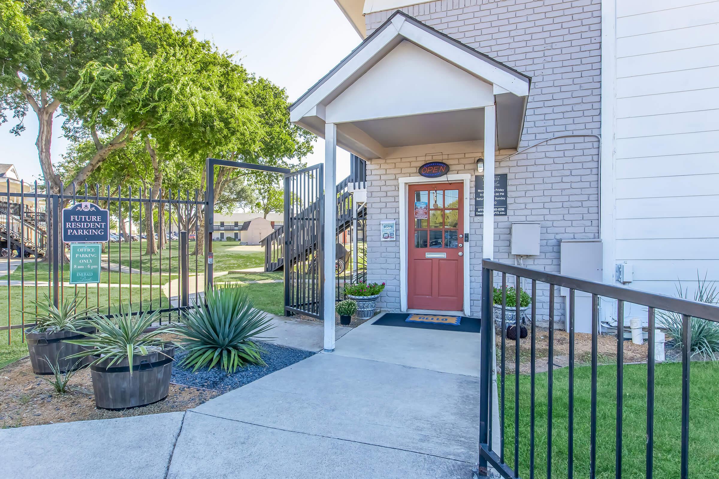 Entrance of a residential building featuring a colorful front door, potted plants, and a gated pathway. A sign indicates future resident parking. Lush green landscaping and nearby stairs leading to upper floors are visible, creating an inviting and well-maintained atmosphere.