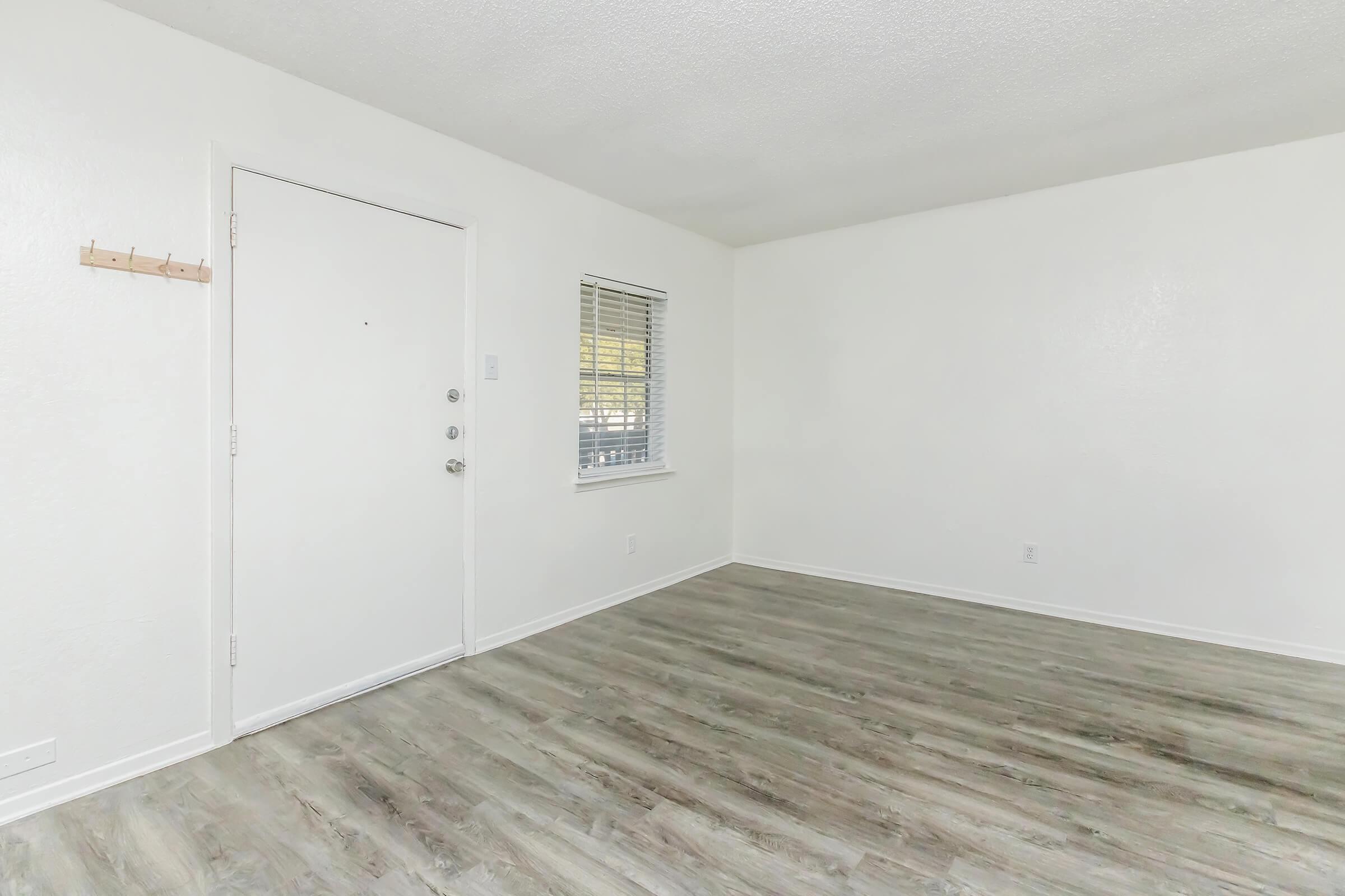 A bright, empty room with white walls and a light-colored wooden floor. A closed door is on the left side, with a small coat hook next to it. A window with blinds allows natural light in, creating an airy atmosphere. The room appears spacious and ready for furnishings.