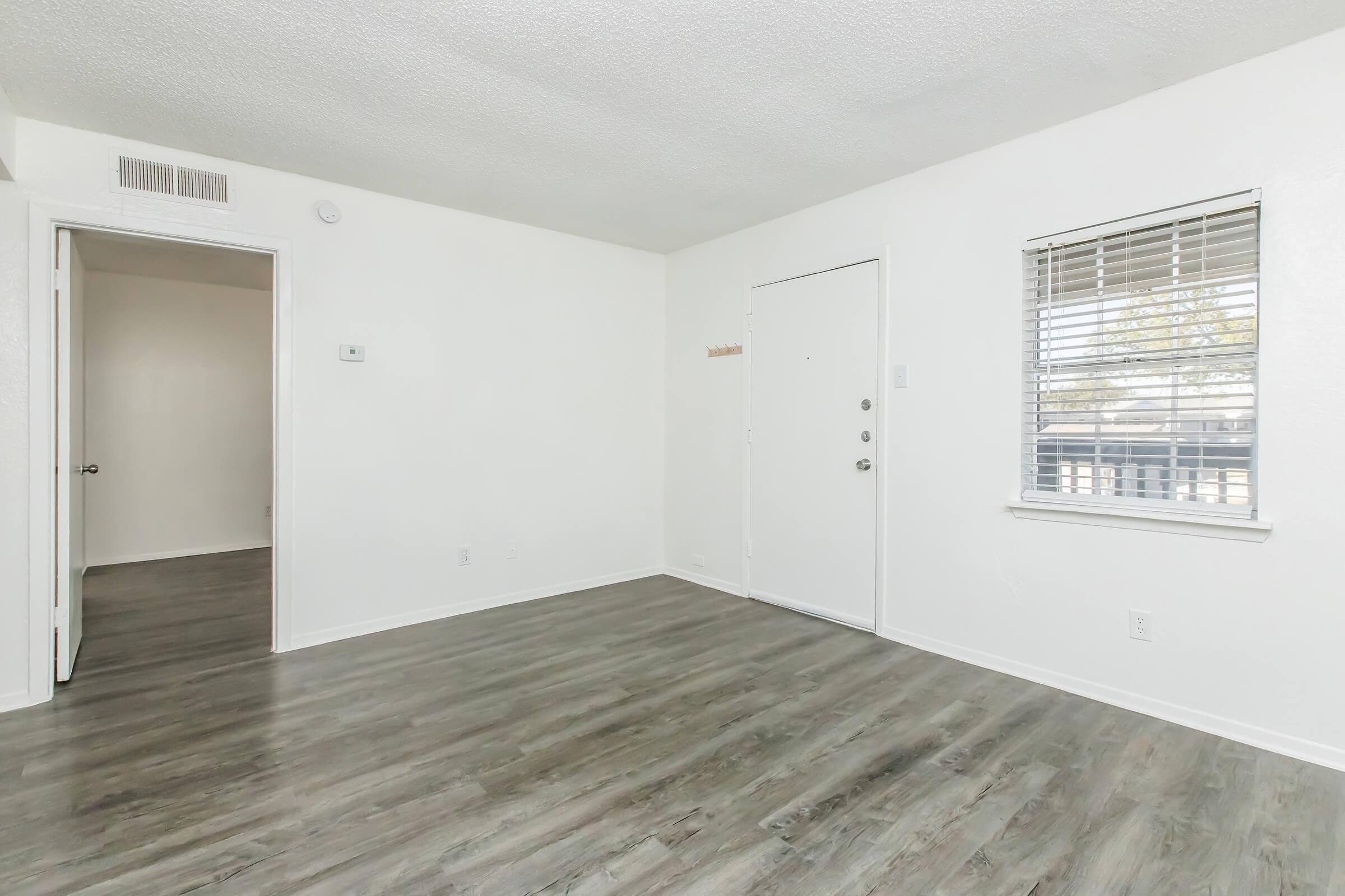 Empty interior of a well-lit room featuring a light-colored wall and wooden flooring. A door leads outside, while a small window with blinds offers natural light. A doorway on the left opens to another room. The space is clean and modern, suitable for various decorating options.