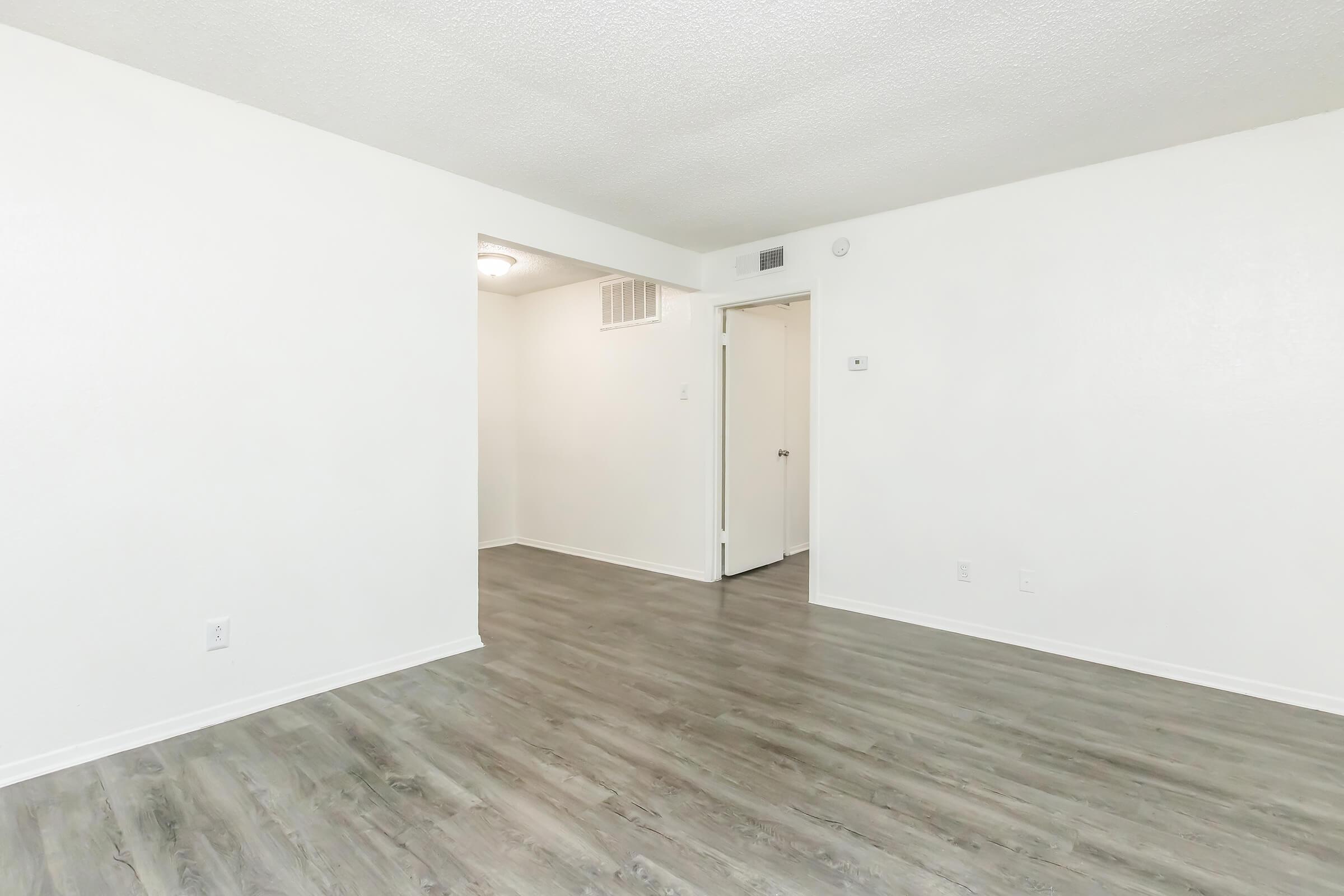 A bright, empty room with light-colored walls and a textured ceiling. The flooring is a light wood finish. An open doorway leads to another area, with a door slightly visible in the background. Natural light illuminates the space, creating a clean and modern aesthetic.