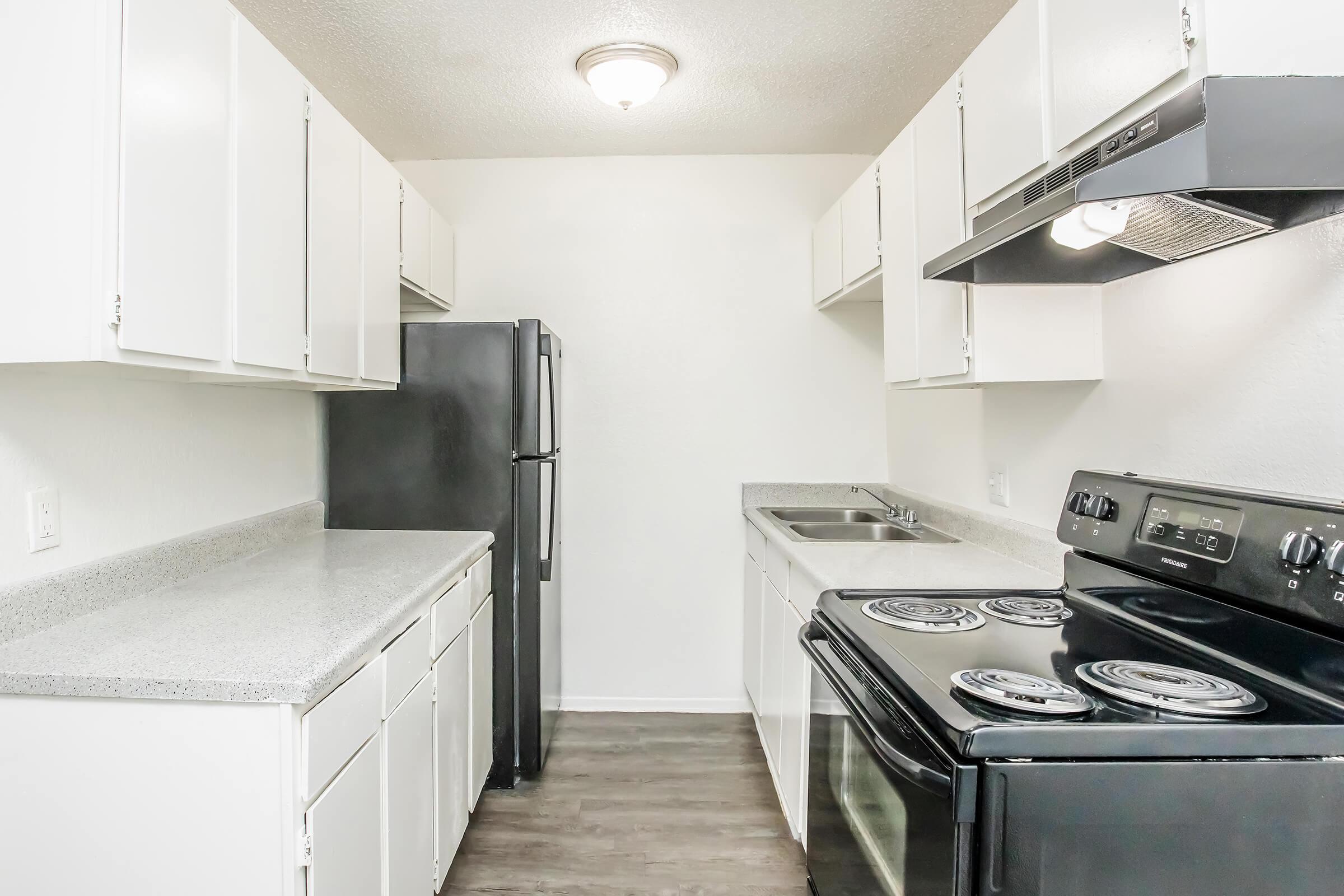 A modern kitchen featuring white cabinets, a black stove, and a refrigerator. The kitchen has gray countertops, a stainless steel sink, and is well-lit by a ceiling fixture. The flooring is a dark wood laminate, creating a clean and inviting atmosphere.