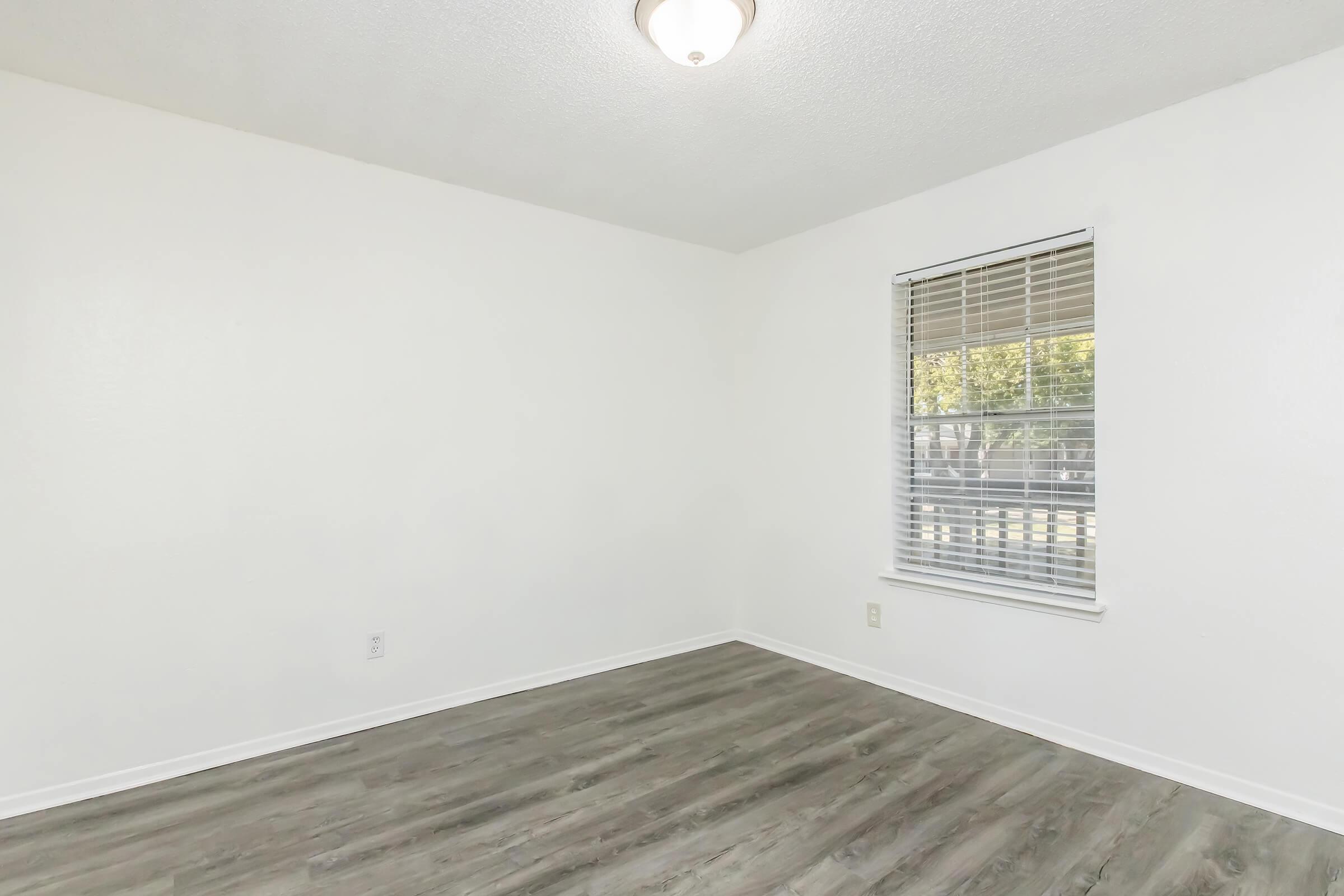 Empty room with white walls and a window featuring blinds. The floor is covered with light-colored wood laminate. A ceiling light fixture is present. The overall appearance is clean and modern, suitable for various uses or decorating.