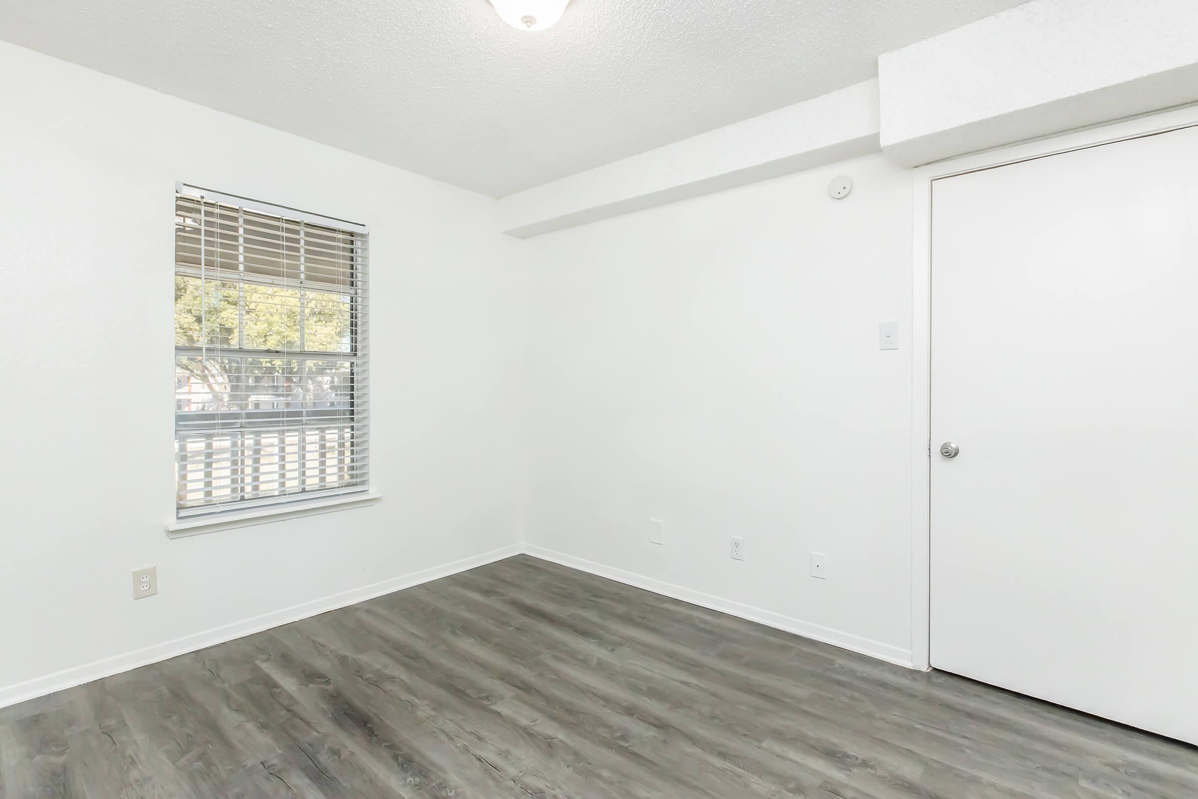 A bright, empty room with light-colored walls and a window letting in natural light. The floor is a modern gray laminate, and there's a closed white door on the right. The room has minimal furniture, creating an open and spacious feel. Ideal for personal customization or decoration.