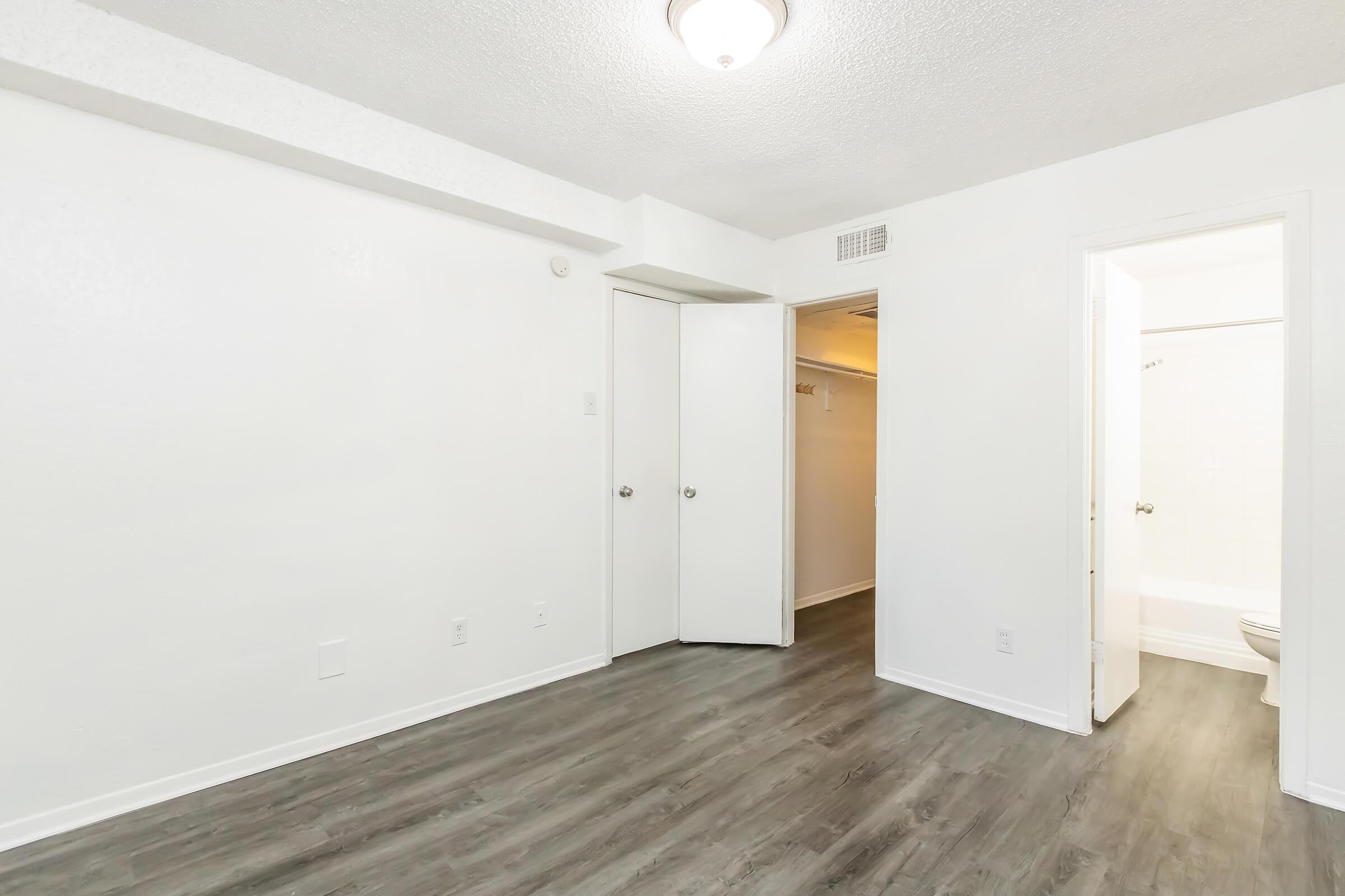 Spacious, empty room with light-colored walls and a modern ceiling light. The floor features a dark laminate finish. There are two doors leading to a closet and a bathroom, with a small window providing natural light. The overall look is clean and minimalistic, suitable for various uses.