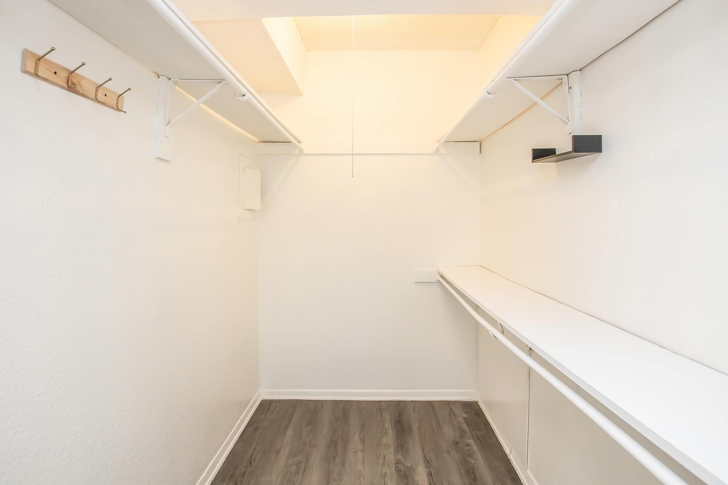 A spacious, empty closet with white walls and a wooden rack on one side. There is a long white shelf running along one wall, and the floor is covered in a dark laminate. The room has a bright light source coming from above, creating a well-lit atmosphere.