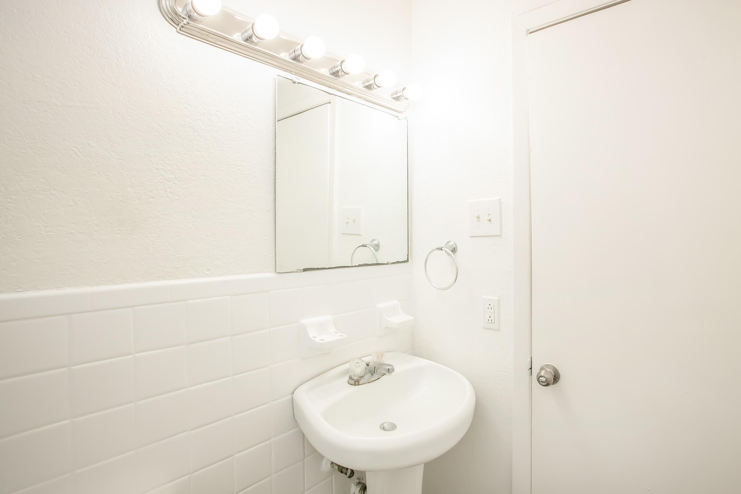 A small bathroom featuring a white wall, a mirror above a sink, a light fixture with multiple bulbs, a hand towel holder, and a closed door. The sink is simple, with a single faucet, and two white soap dishes are mounted on the wall above it. The overall aesthetic is clean and minimalistic.