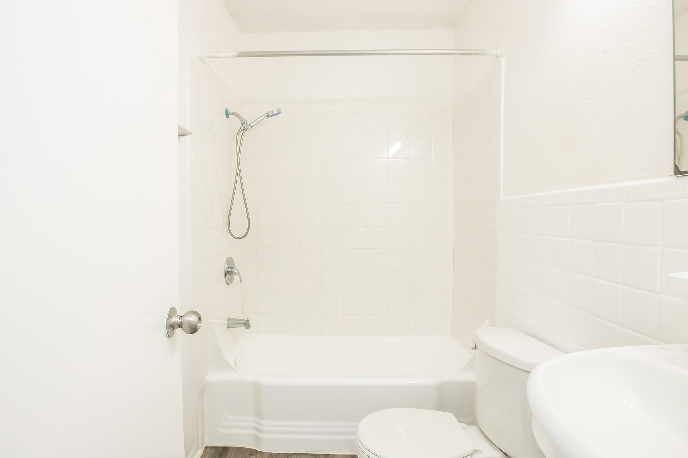 A clean, white bathroom featuring a bathtub with a showerhead, a toilet, and a sink. The walls are tiled with white tiles, and the floor has a wooden finish. Natural light illuminates the space, creating a bright and airy atmosphere.
