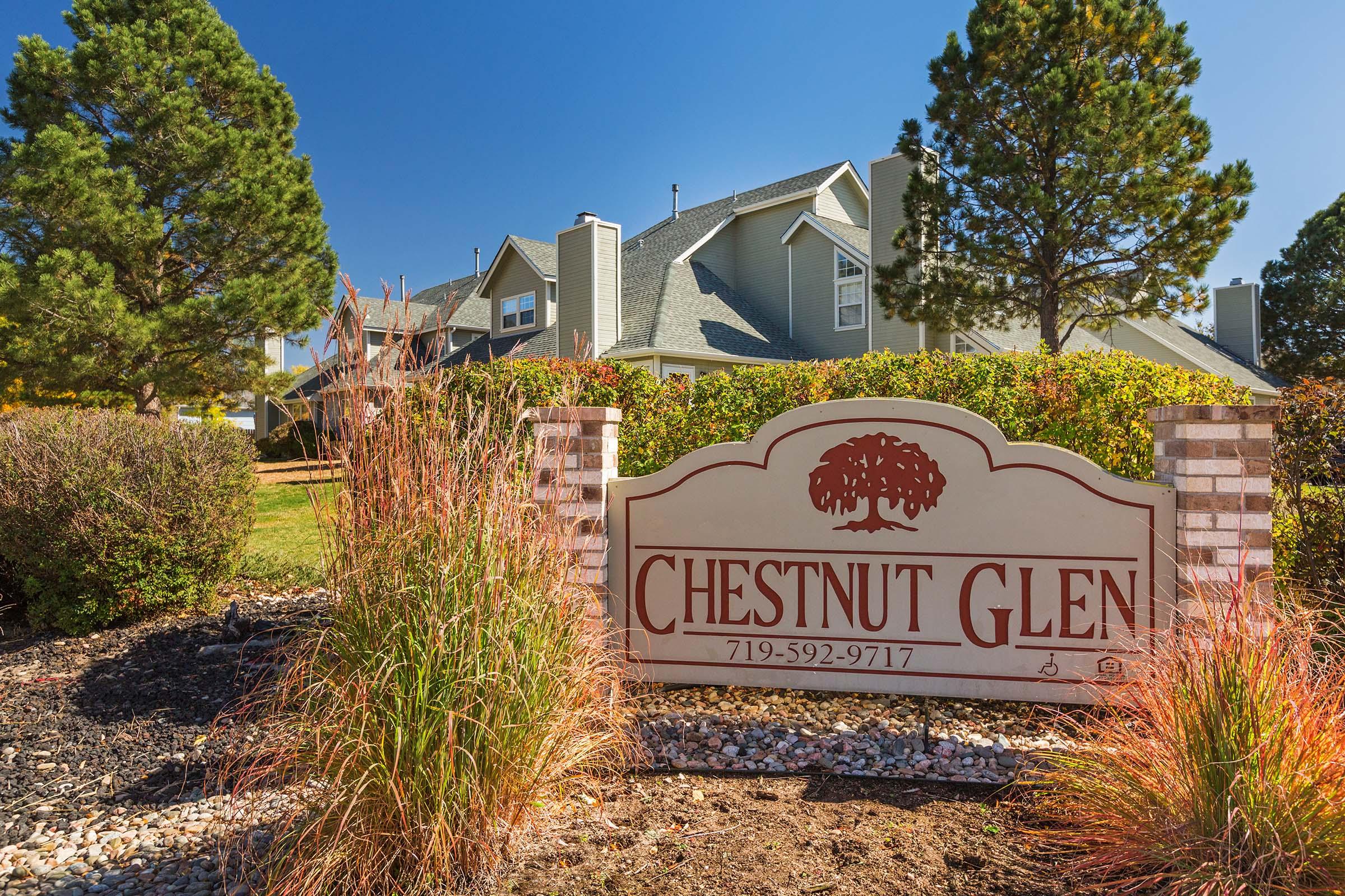 A welcoming sign for "Chestnut Glen" surrounded by neatly trimmed bushes and ornamental grasses. In the background, a residential building with a peaked roof is visible under a clear blue sky.