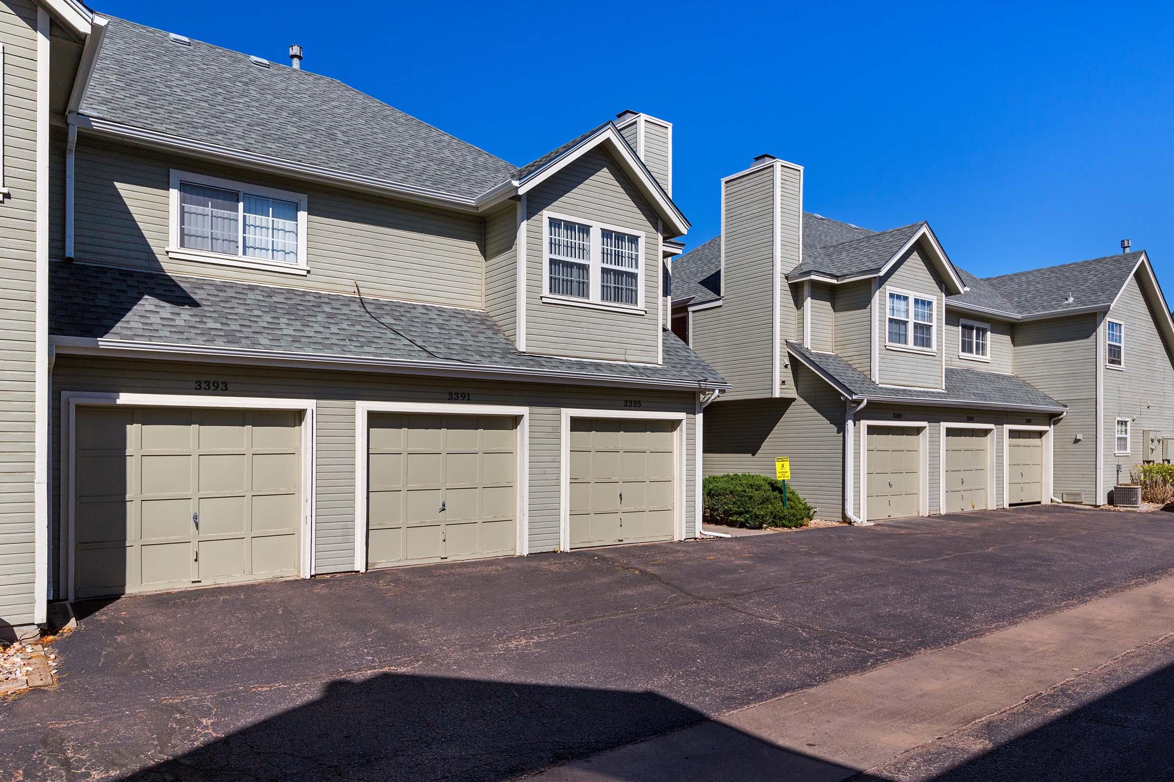 A row of townhouses featuring beige exteriors and grey roofs, each with a garage door. The sky is clear blue, and the pavement in front is dark asphalt. The setting appears well-maintained with shrubs along the side.