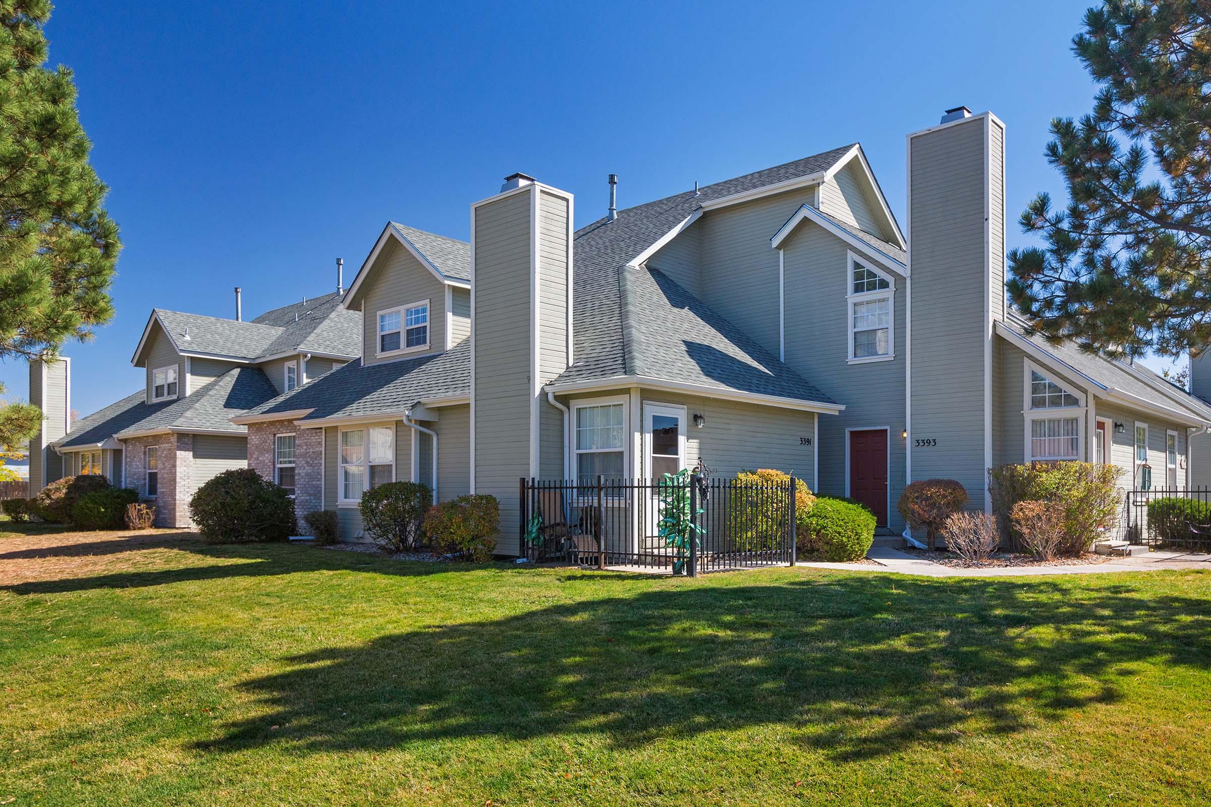 A sunny view of a two-story residential building with a sloped roof, surrounded by well-maintained grass and landscaping. The building features multiple windows, a fenced patio area, and tall trees in the background, indicating a peaceful neighborhood setting.