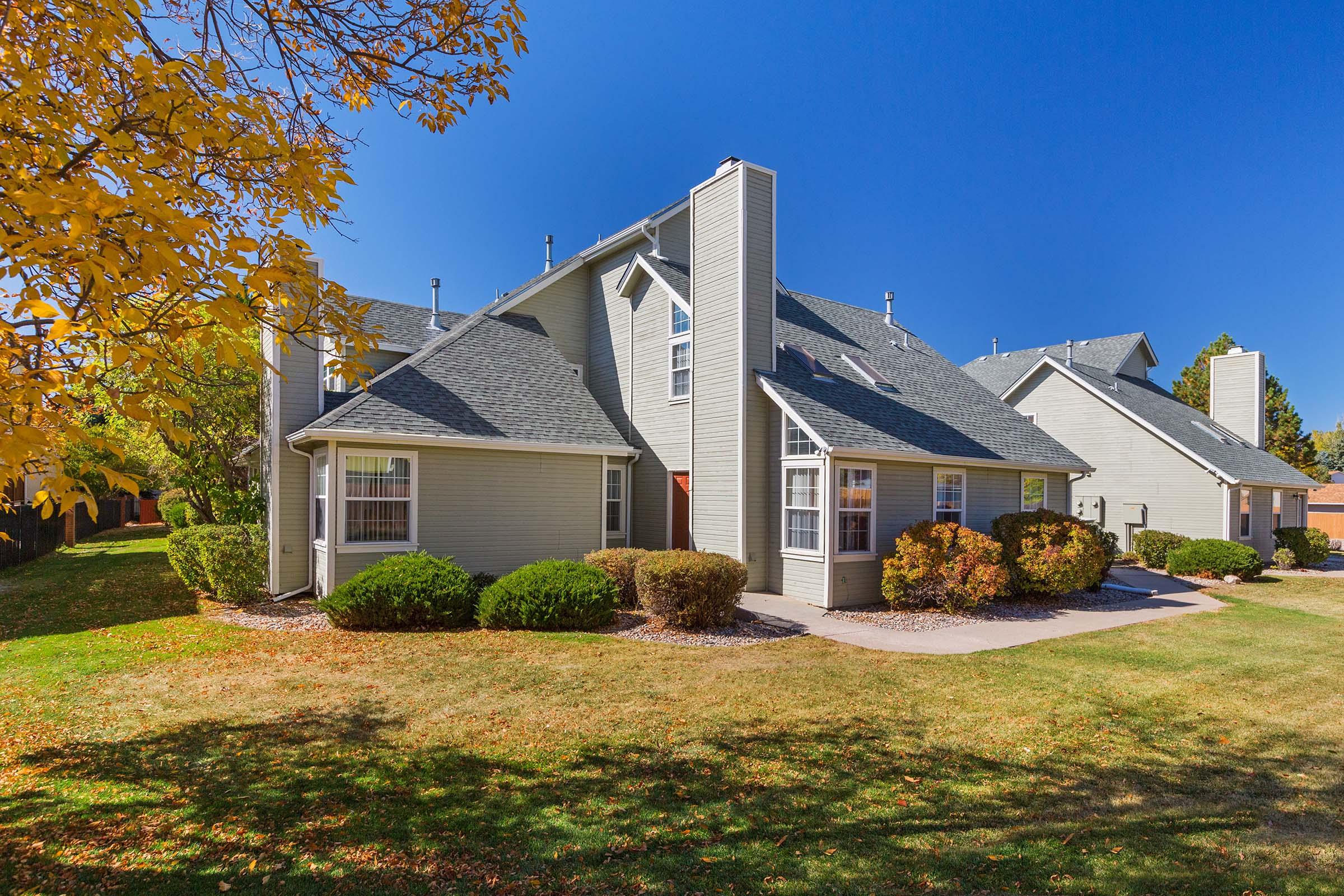 A well-maintained, two-story house with a gray roof and beige siding, surrounded by manicured lawns and colorful autumn foliage. The clear blue sky provides a bright backdrop, highlighting the architectural features of the home and the lush landscaping.